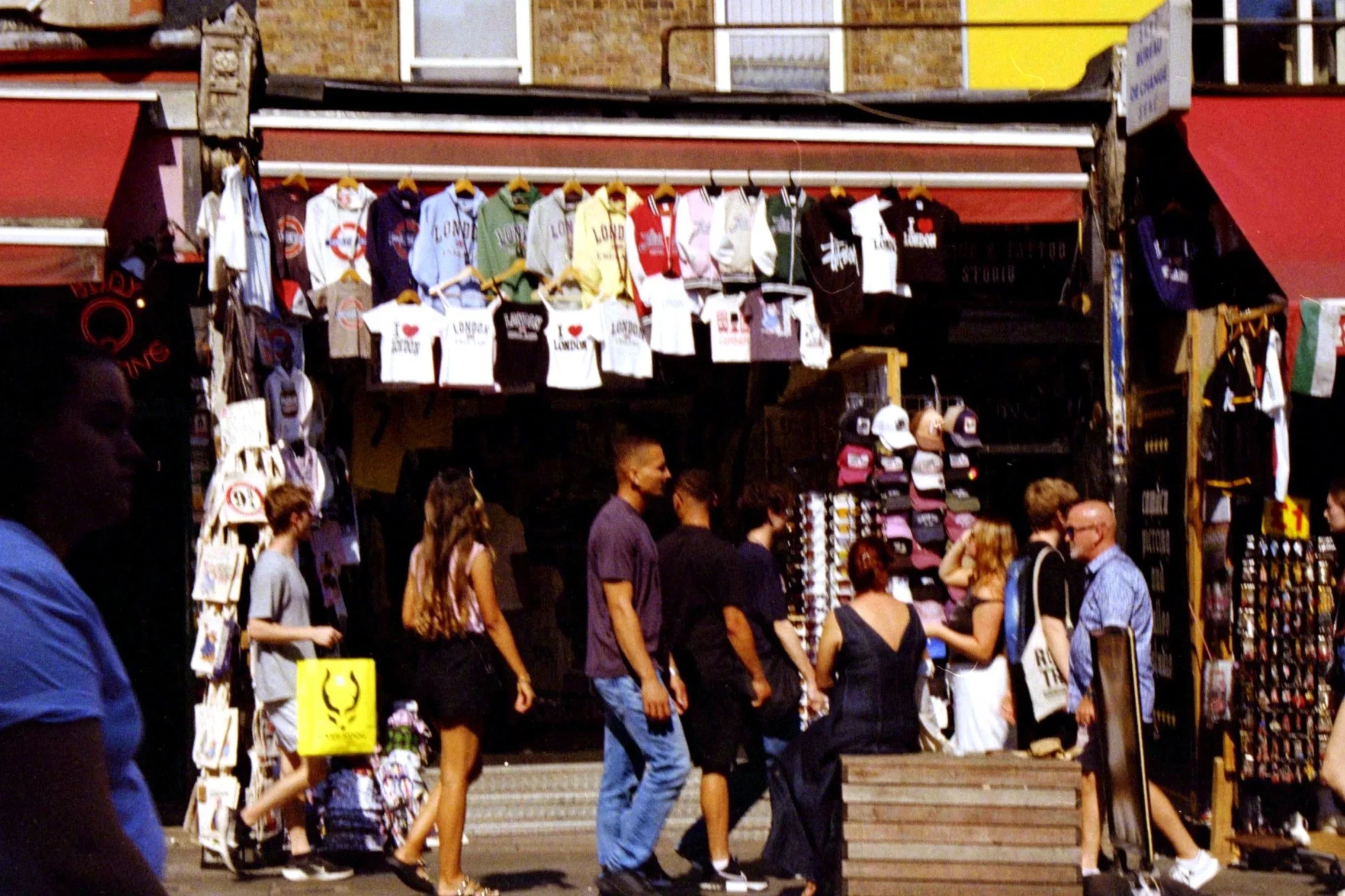 A crowded outdoor market stall sells various clothing items including shirts, hoodies, and caps, with people browsing and shopping in front of it.