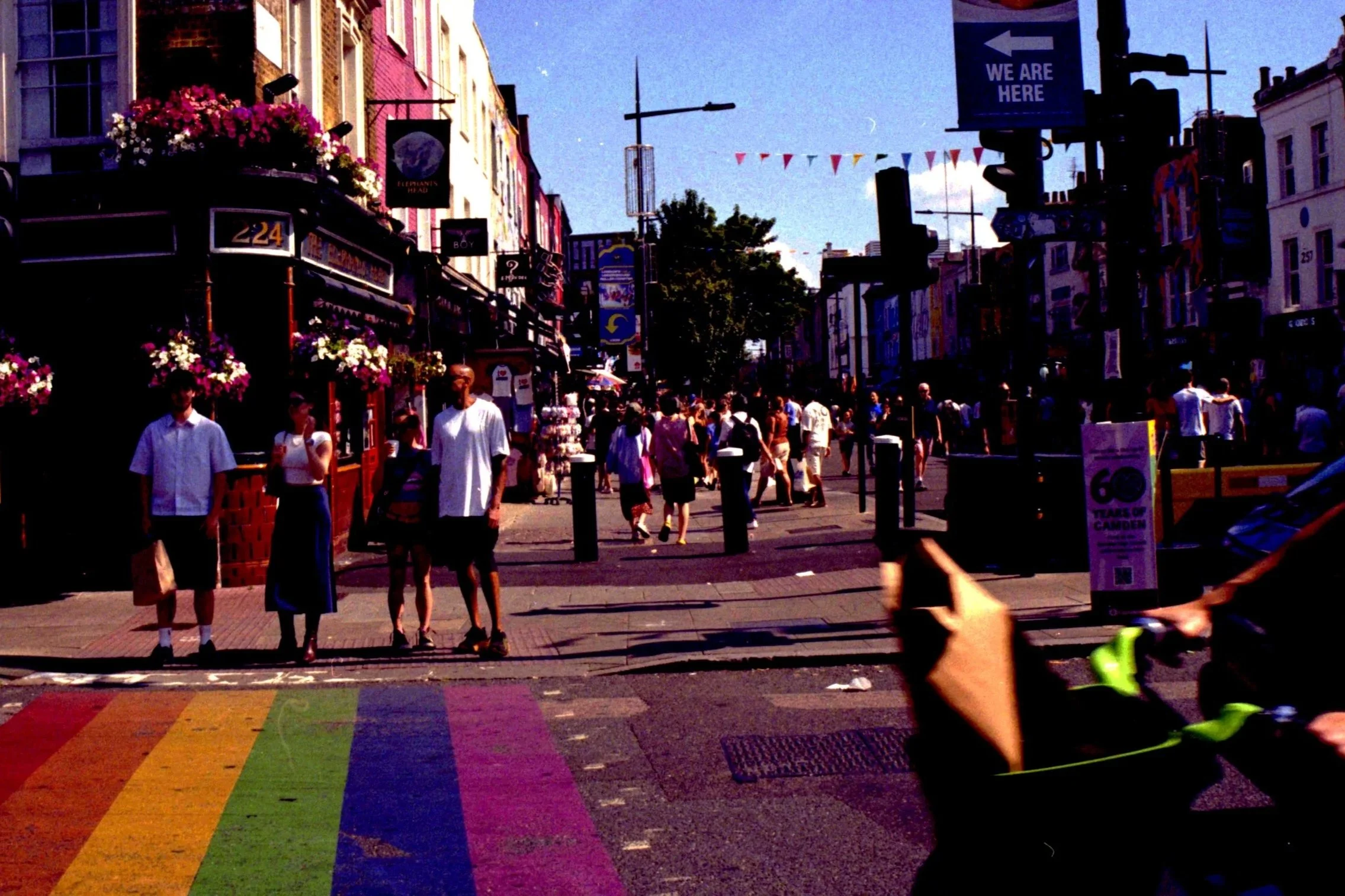 A busy city street scene with people walking, storefronts, street signs, and a rainbow crosswalk on the pavement.