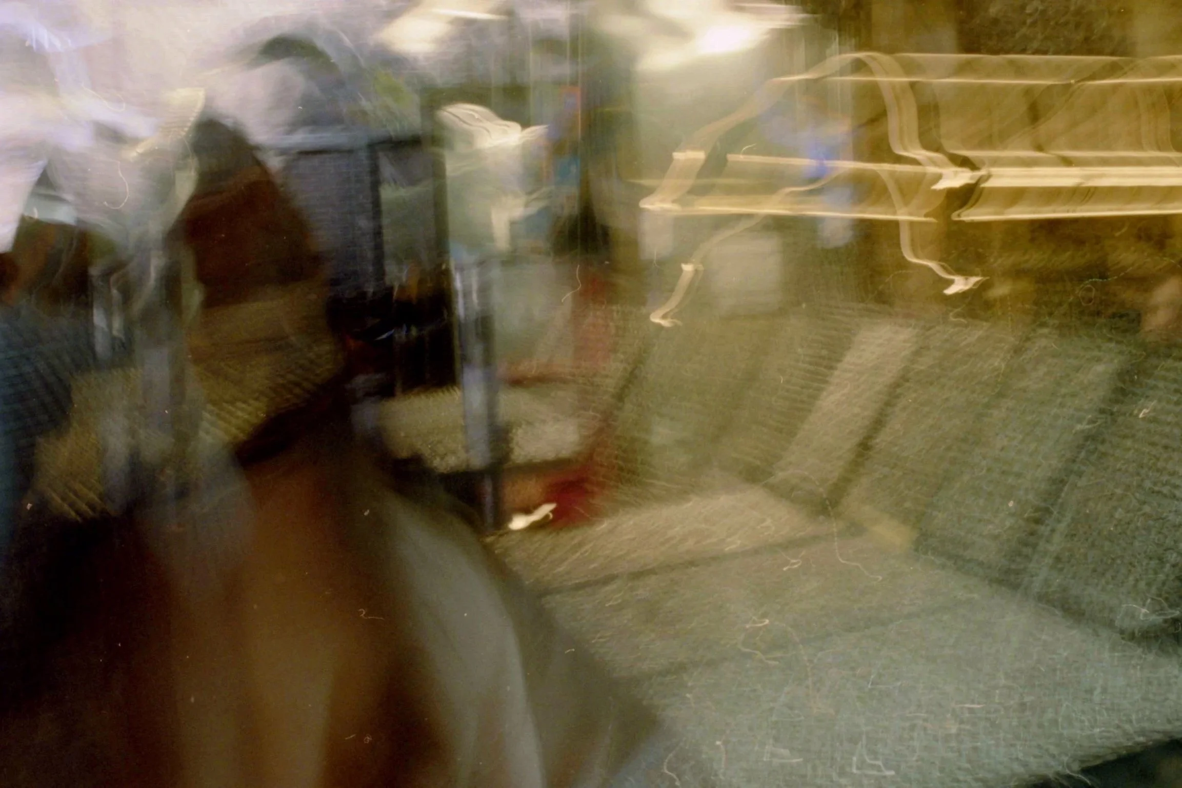 The image appears blurry and distorted with streaks of light, showing a reflection of a person taking a photo through a glass window or door at night. Some outdoor elements, possibly chairs or tables, are faintly visible in the background.