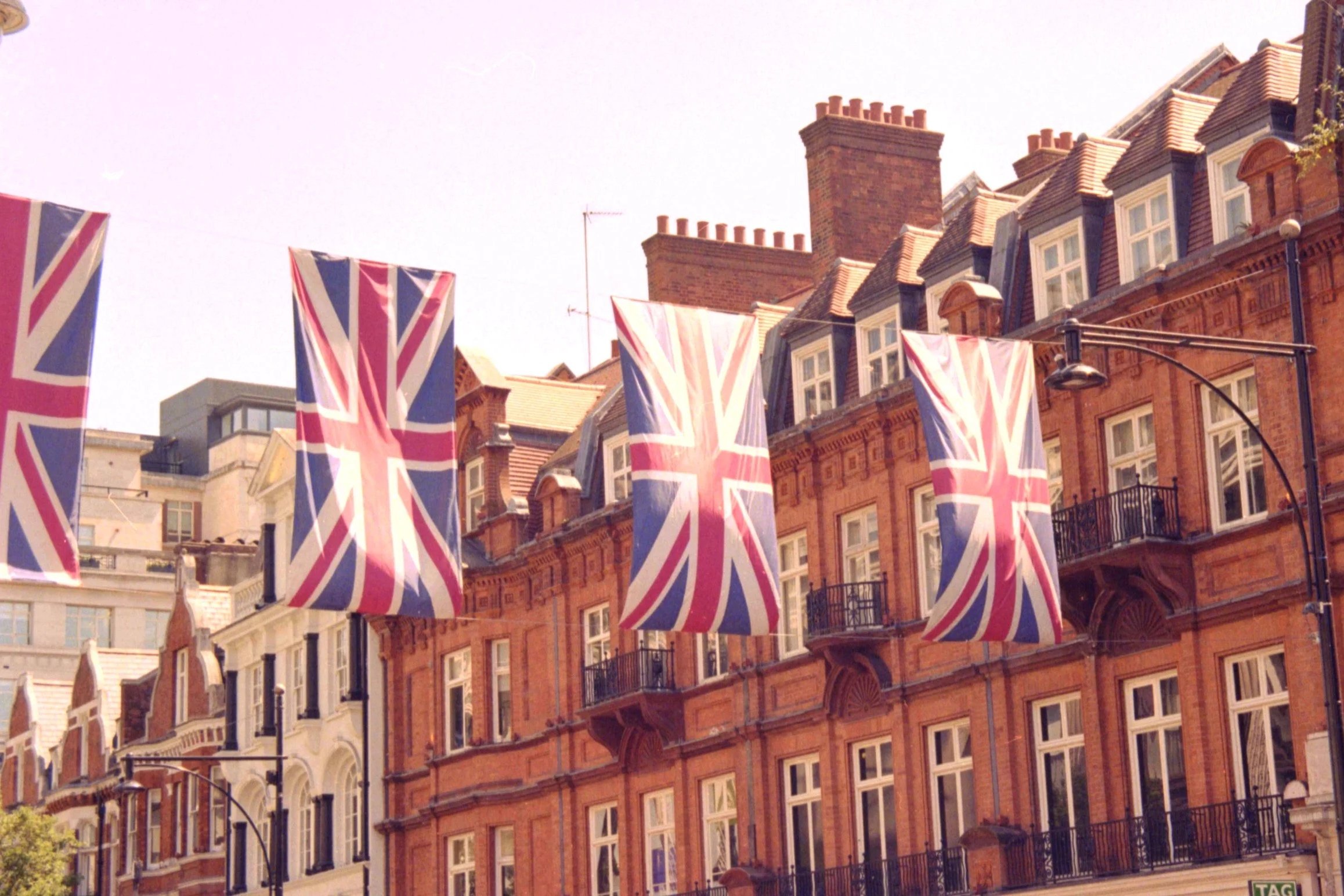 Multiple British flags hanging in front of historic red brick buildings on a city street.