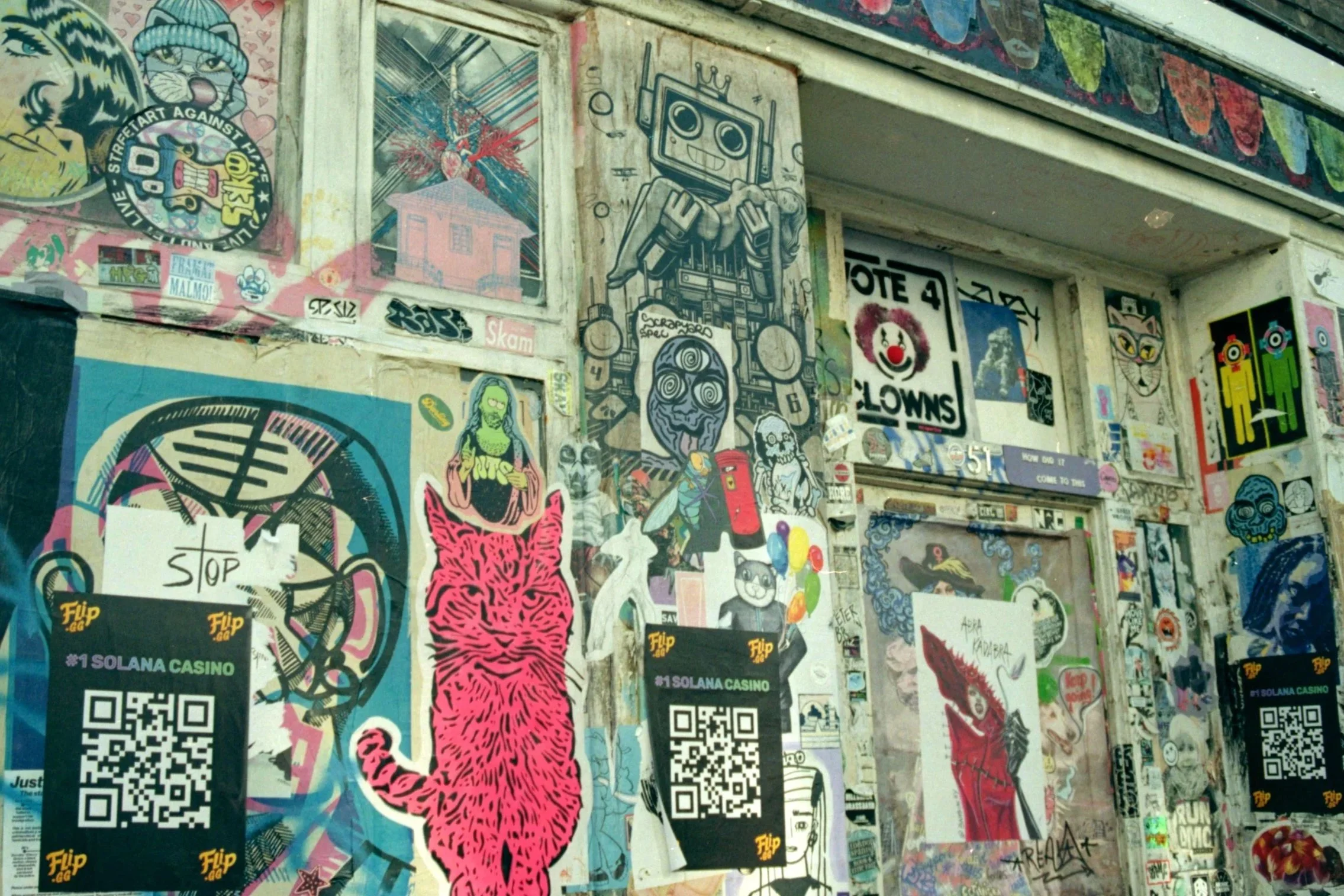 A wall covered with various pieces of graffiti art, stickers, posters, and QR codes, including a cartoon girl with pink tiger print clothing, a colorful clown face, and a boombox with spiral eyes.