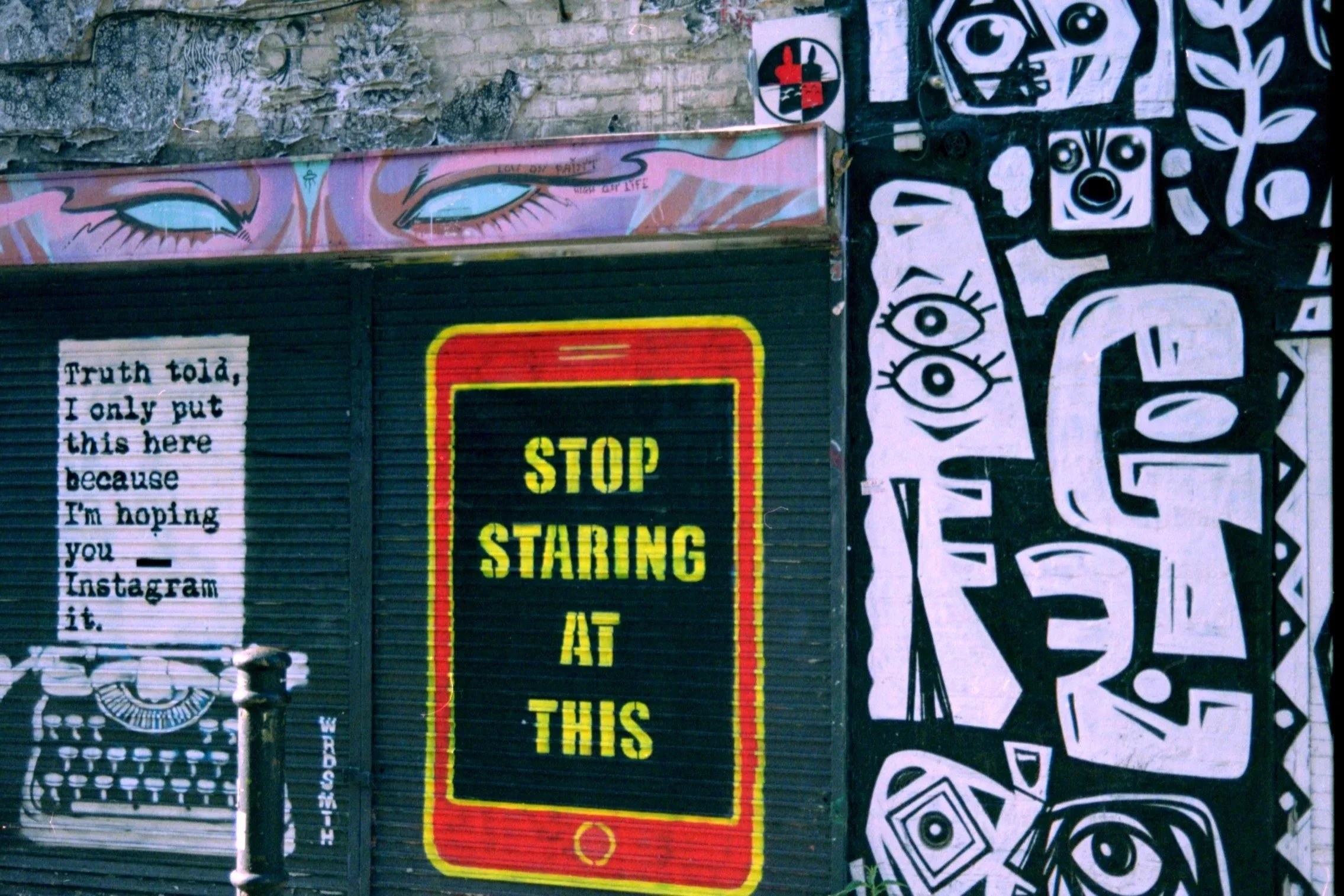 Street mural artwork featuring stylized eyes, a patterned face, and text including "Truth told, I only put this here because I'm hoping you Instagram it" and a yellow and red "STOP STARTING AT THIS" sign.