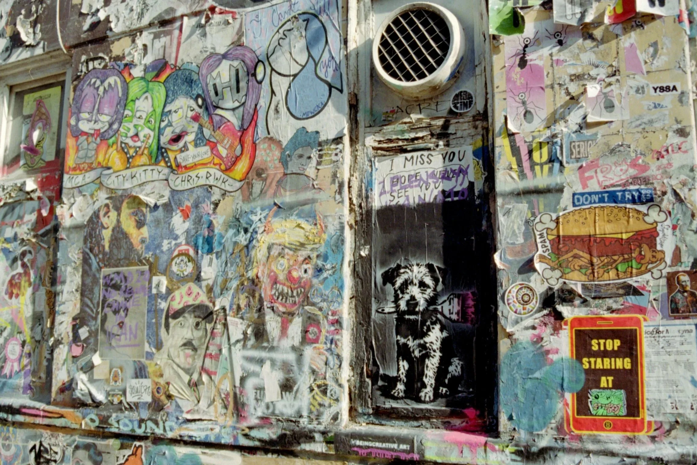 A collage of street art, stickers, and posters on a worn, peeling wall with graffiti and drawings, including abstract images, text, and characters.