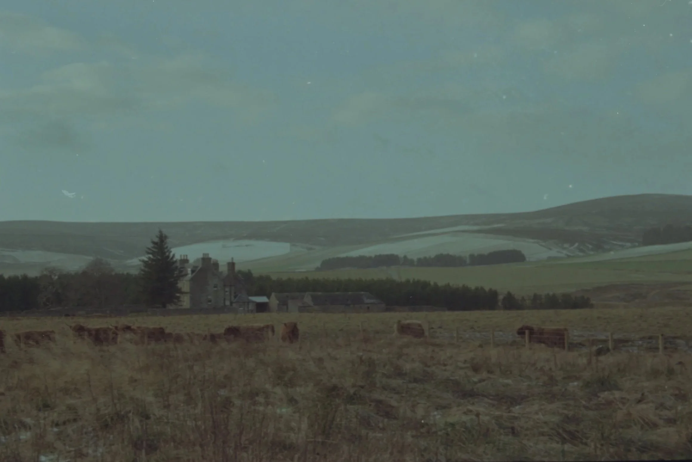 A rural landscape with rolling green hills, a lake, and some houses in the foreground, under a cloudy sky.