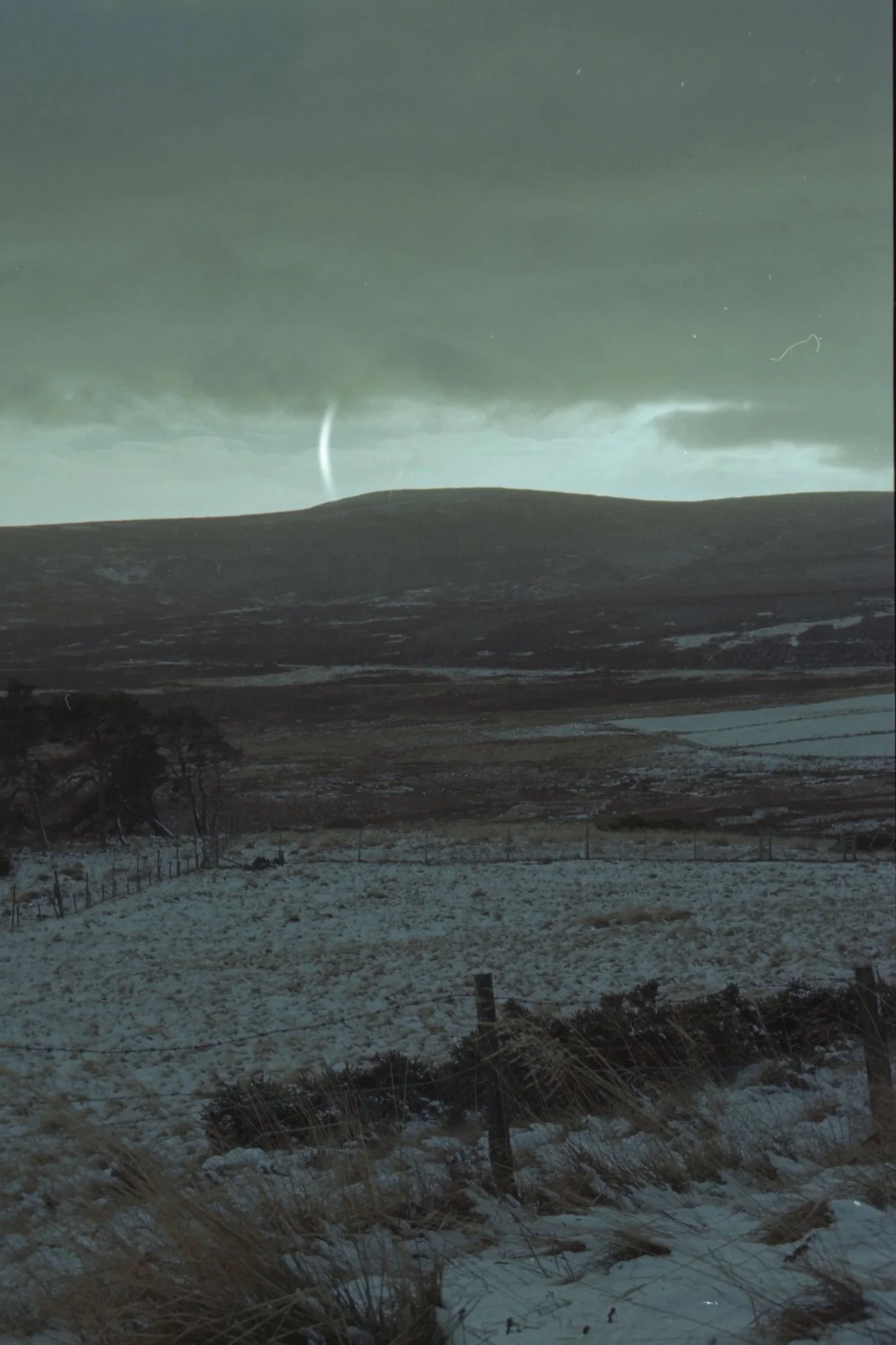 Snow-covered field with a fence, trees, a distant hill, dark cloudy sky, and a lightning bolt.