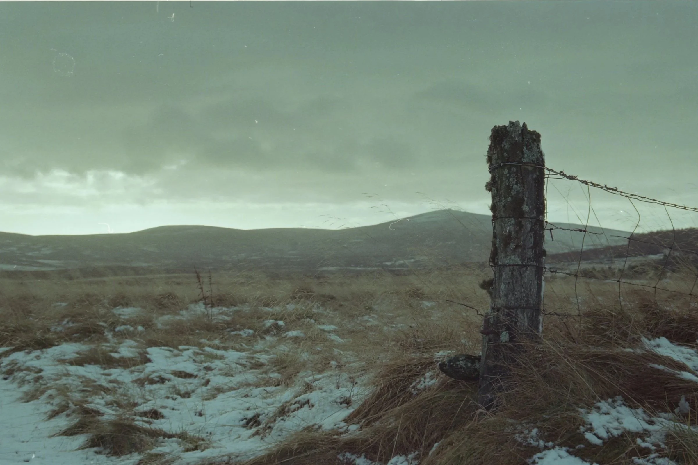 A weathered wooden fence post with barbed wire stands in a rural landscape with snow-covered ground and grassy patches, under a cloudy sky.