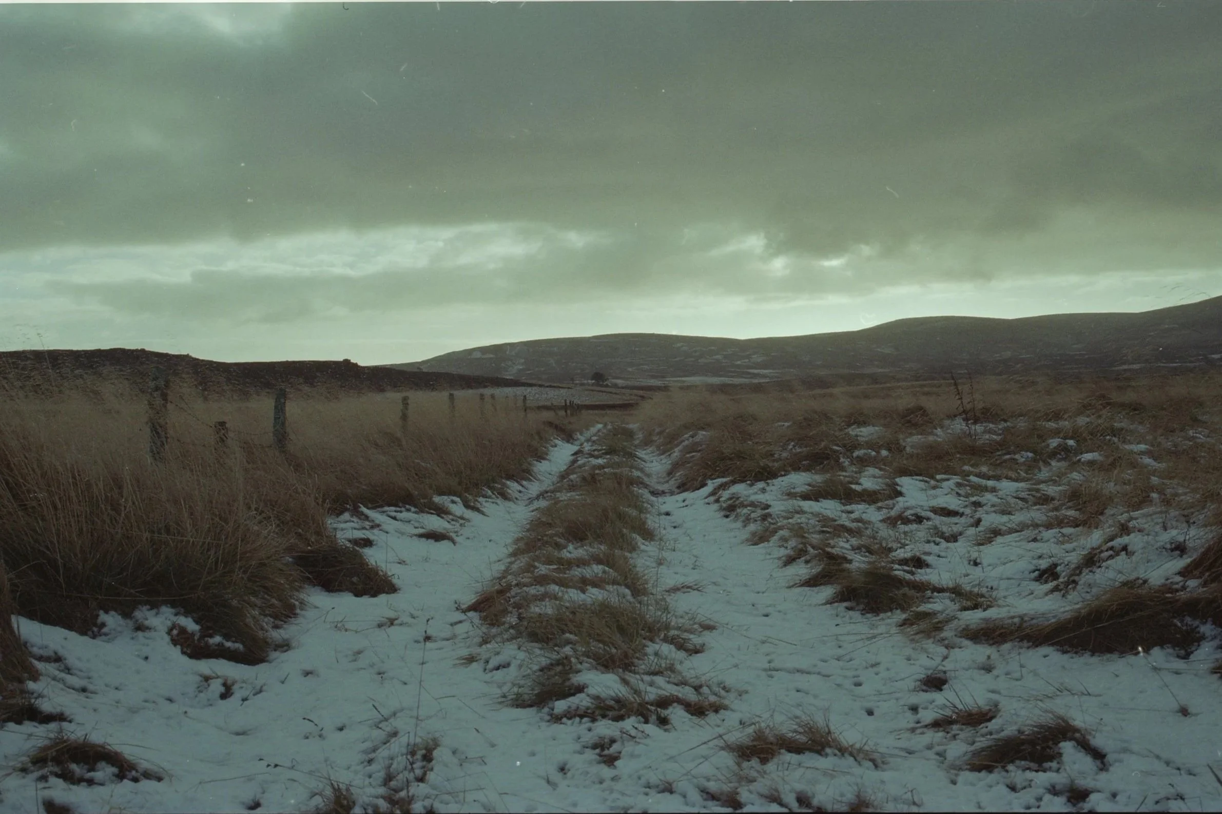 A snow-covered dirt path in a hilly landscape with dry grass and a cloudy sky.