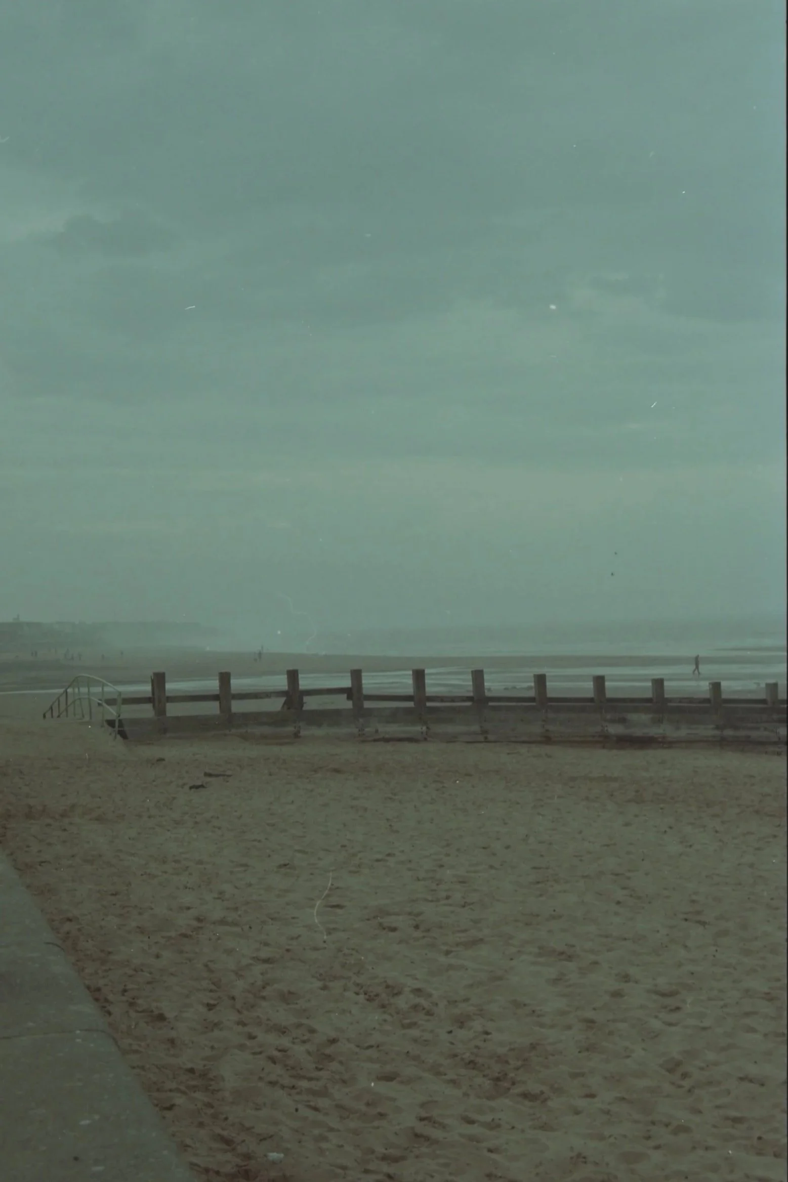 A sandy beach with a boardwalk and railing, with the ocean and cloudy sky in the background. A person is walking along the shore.