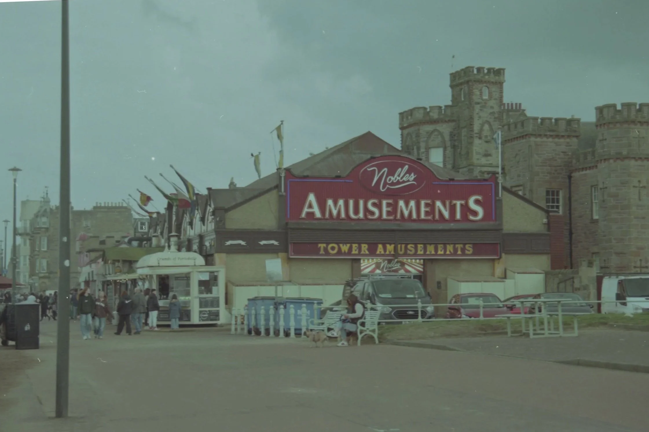 A street scene with a building having a sign that reads 'Nobles Amusements' and 'Tower Amusements'. There are people walking along the sidewalk, parked cars, and flags waving in front of the building. A castle-like structure is visible in the backgro