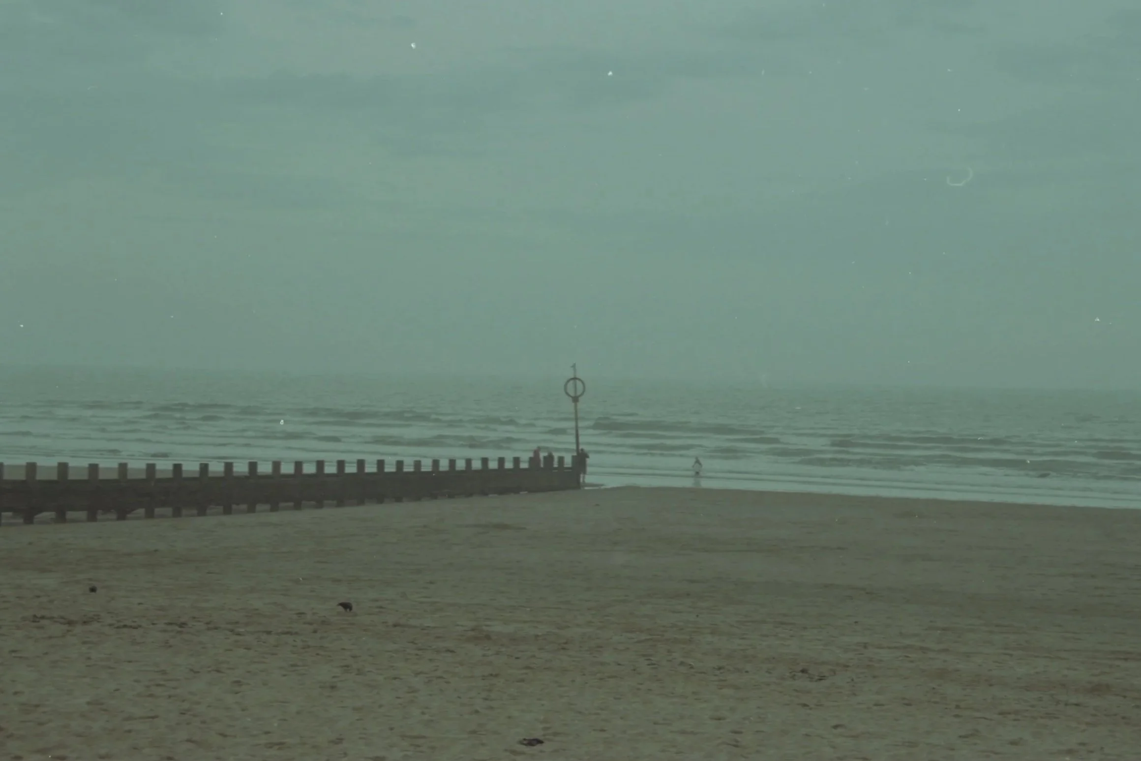 A seaside scene with a beach, pier, and a person walking near the water under an overcast sky.