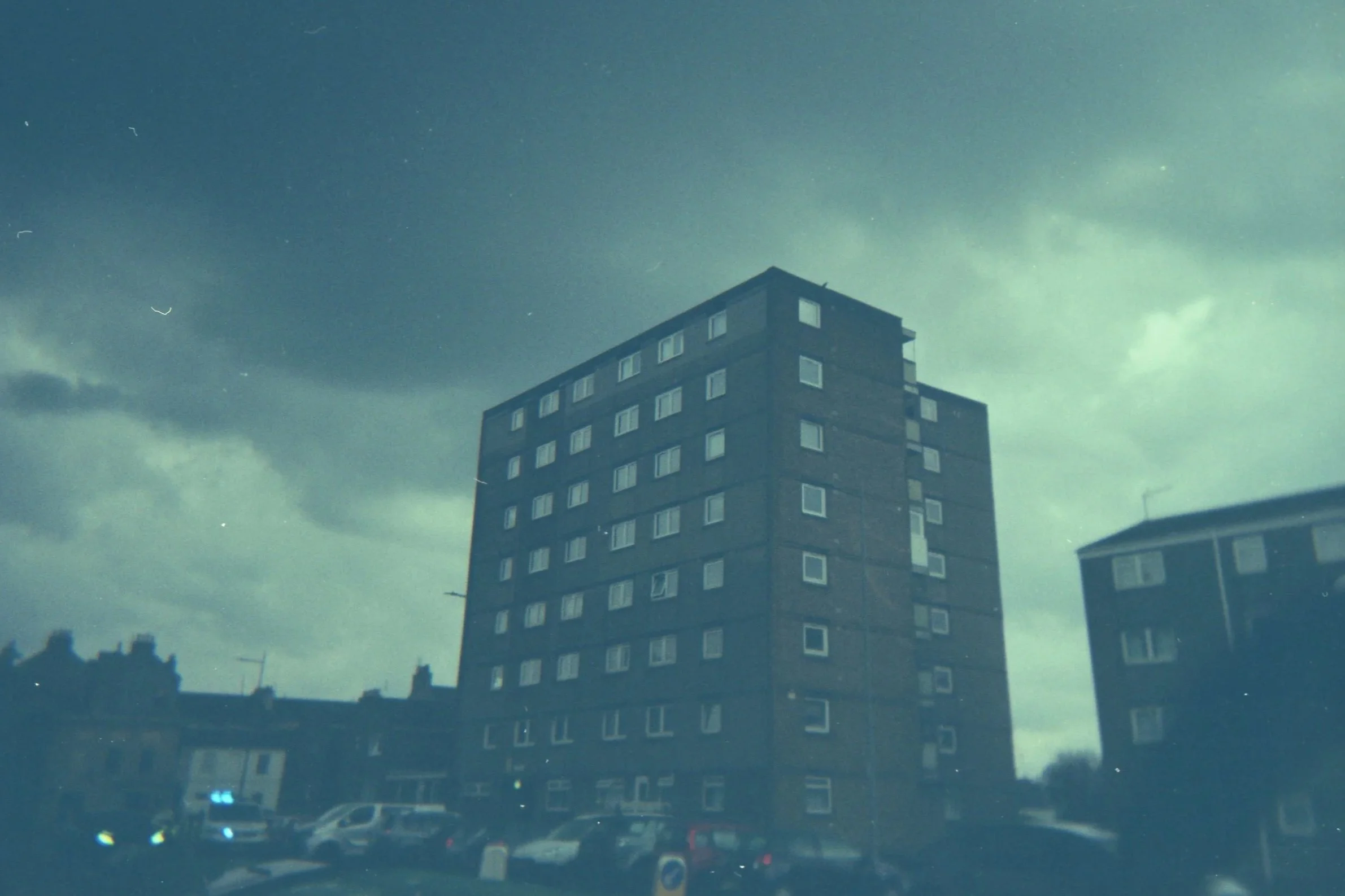 A tall, brick apartment building with multiple windows is seen against a dark, cloudy sky.