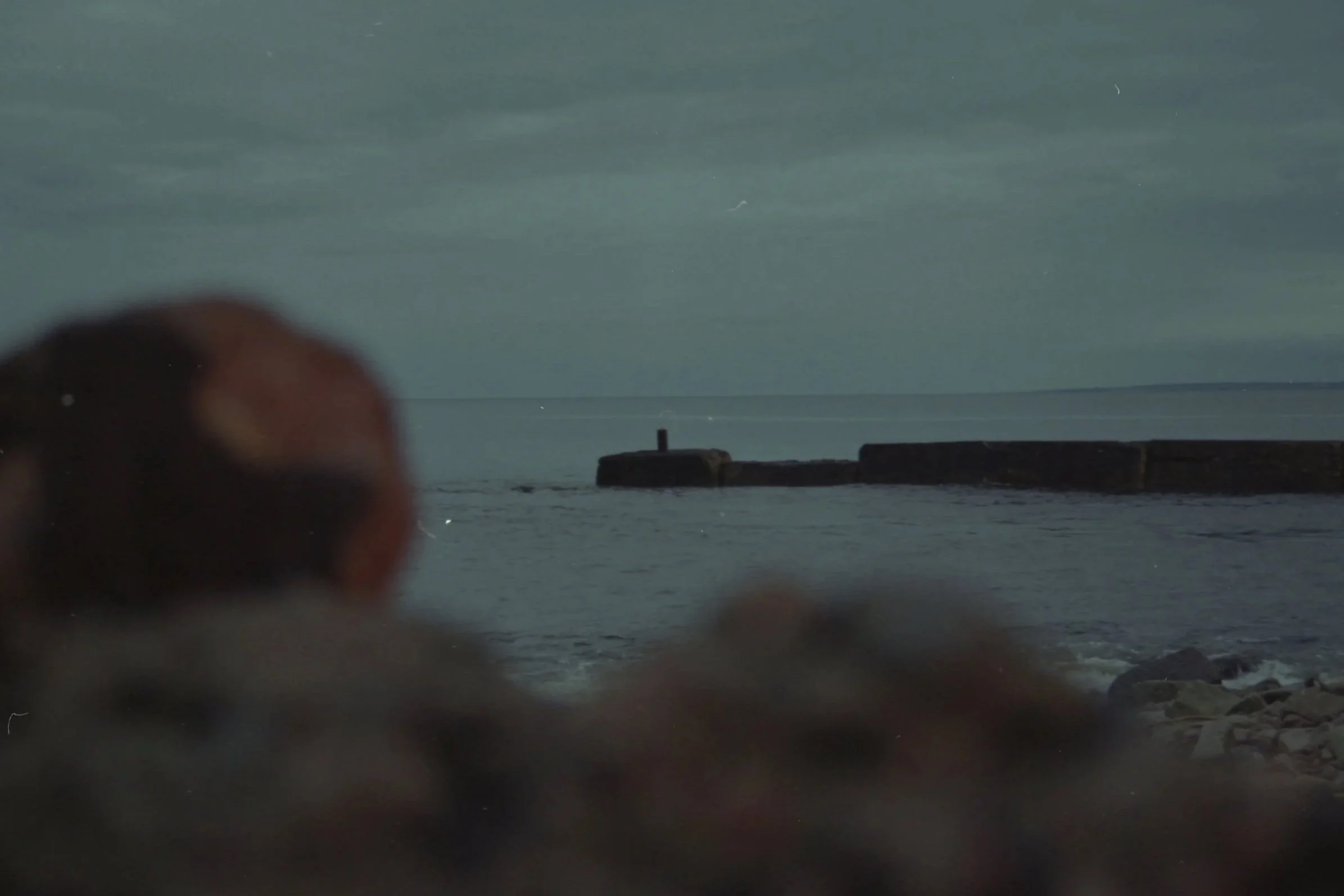 A blurry image showing a body of water with a breakwater or pier structure extending into the water, under a cloudy sky with some streaks of light or snow.