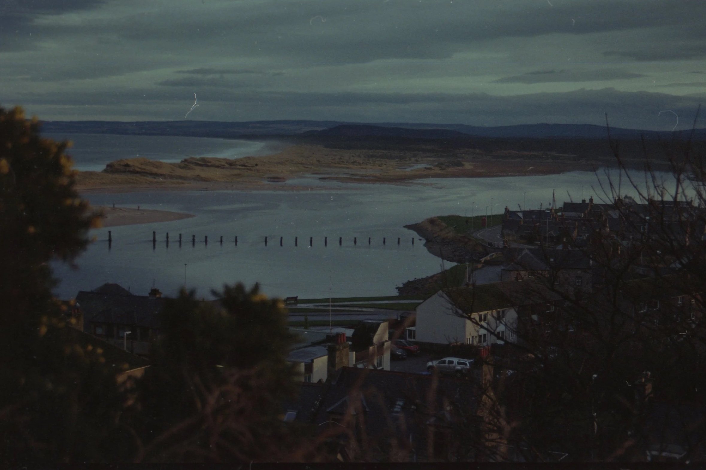 Nighttime scene of a body of water with small houses along the shore, buildings, trees, hills in the distance, and cloudy sky overhead.