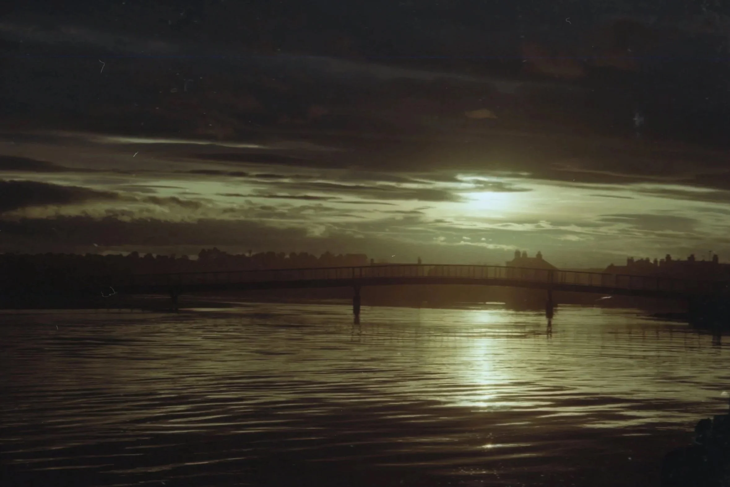 Nighttime scene of a body of water with a bridge in the background, lit partially by moonlight through clouds, with reflections on the water's surface.