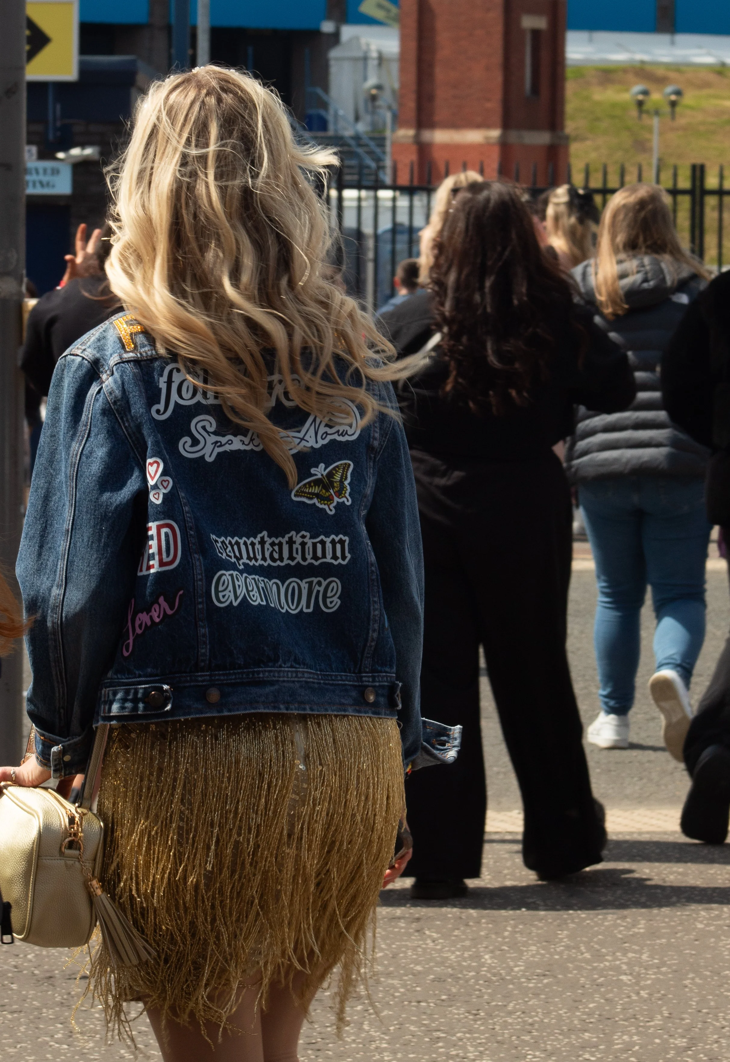 A woman with curly blonde hair wearing a decorated denim jacket and shiny gold fringe skirt, carrying a cream-colored handbag, standing among a group of people outdoors.
