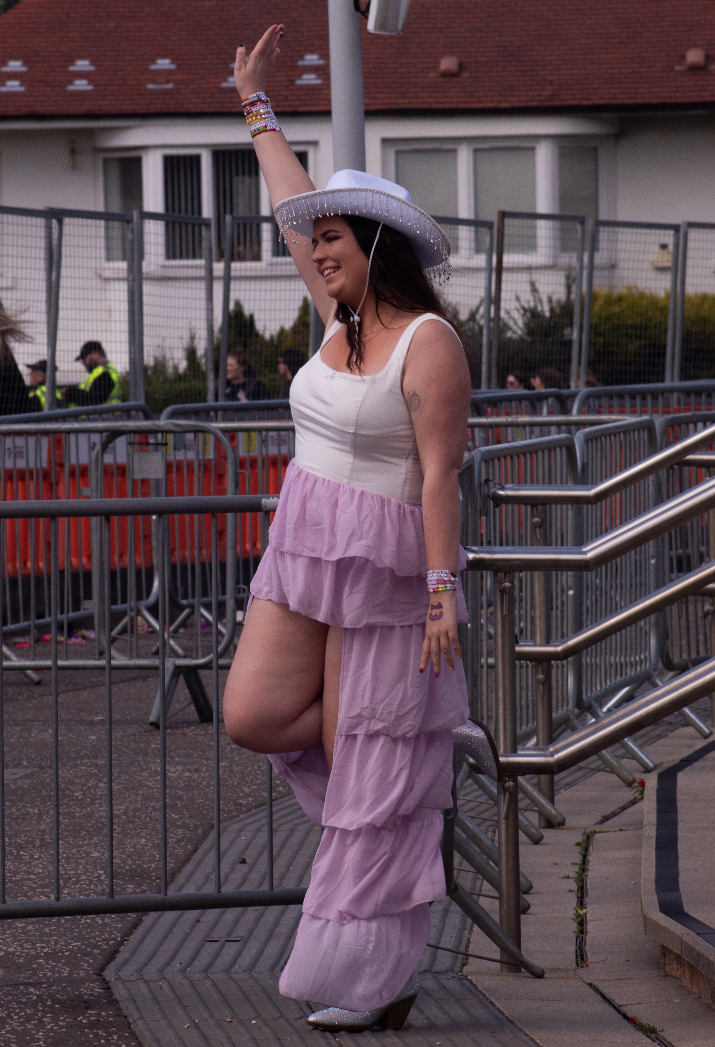 Young woman wearing a white wide-brimmed hat decorated with pearl-like beads, a sleeveless cream top, layered pink skirt, and shiny silver high heels, smiling and raising her arm, standing behind metal barricades on a city sidewalk near a building wi