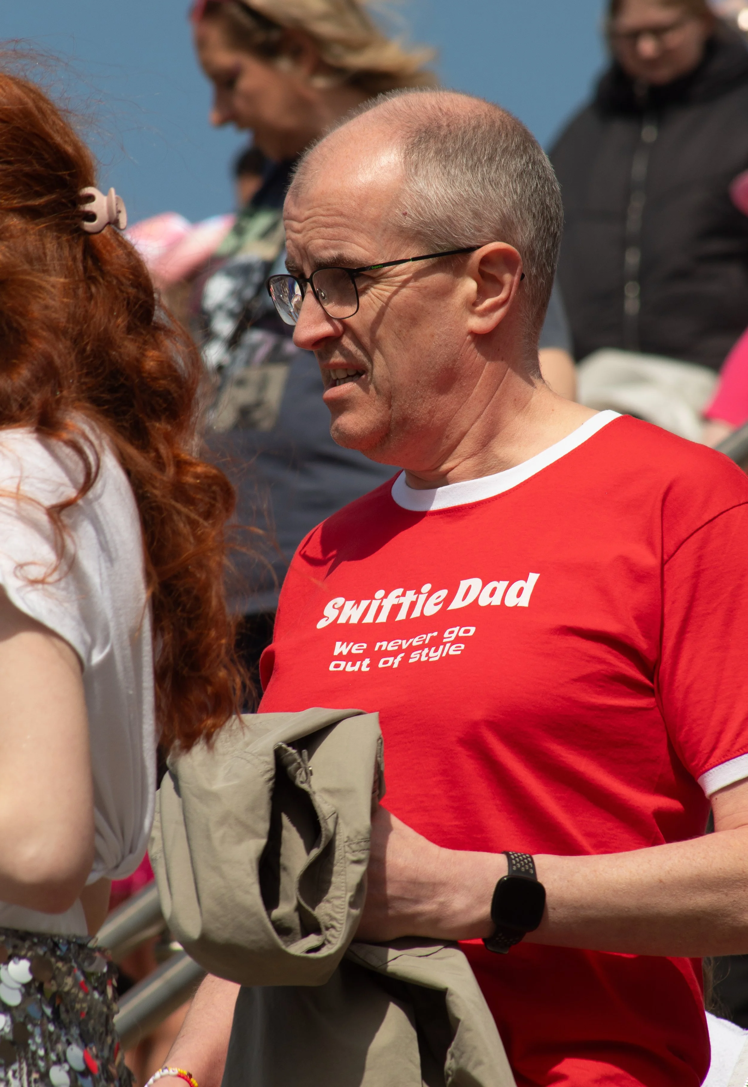 A man with short gray hair, wearing glasses and a red t-shirt with the words 'Swiftie Dad' and 'we never go out of style' on it, is talking to a woman with curly red hair, who is also wearing a white top. They appear to be at an outdoor event with ot