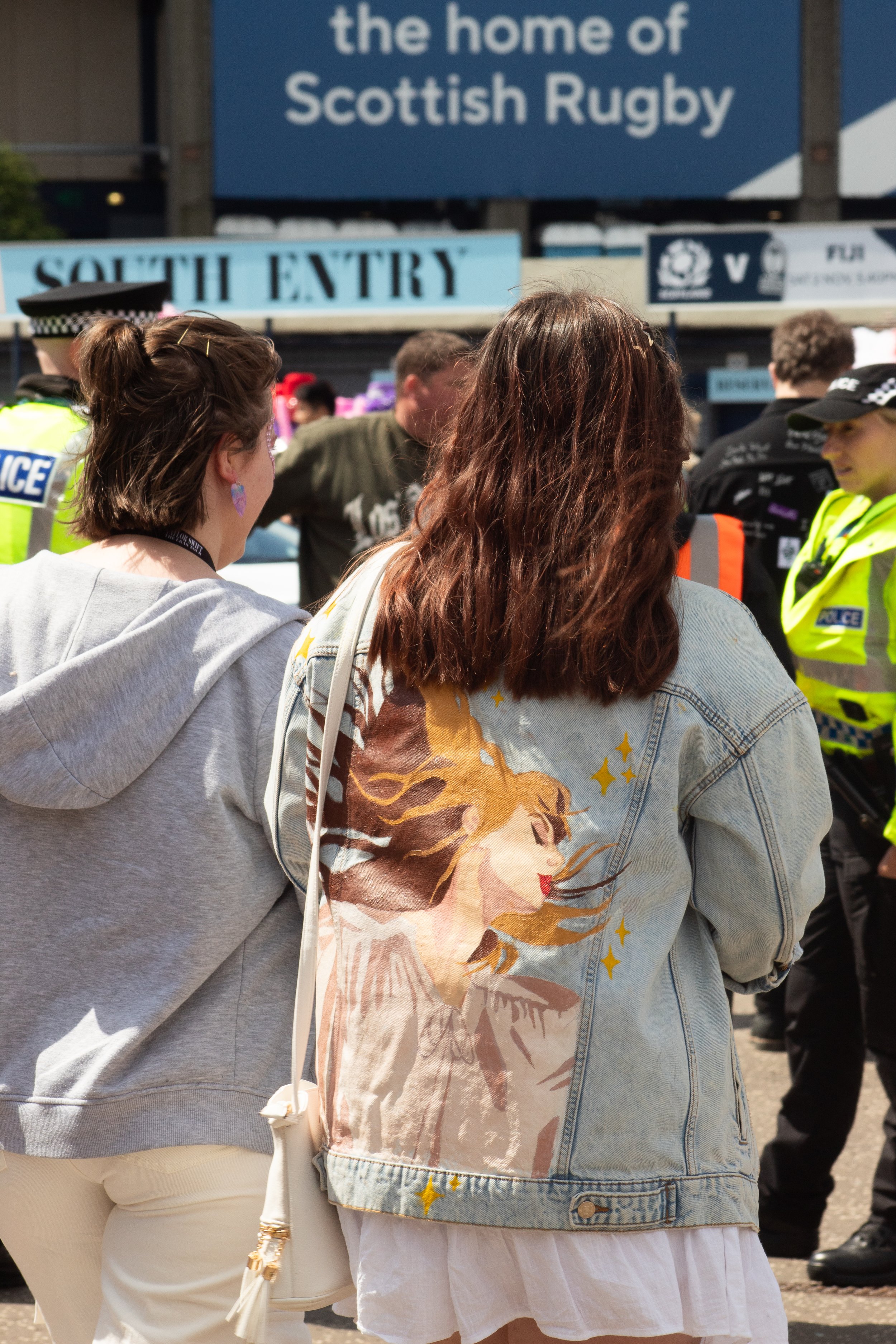 People gathered outside near a sign that reads 'the home of Scottish Rugby,' with some police officers in high-visibility jackets and others in casual clothing, and a blue 'South Entry' sign in the background.