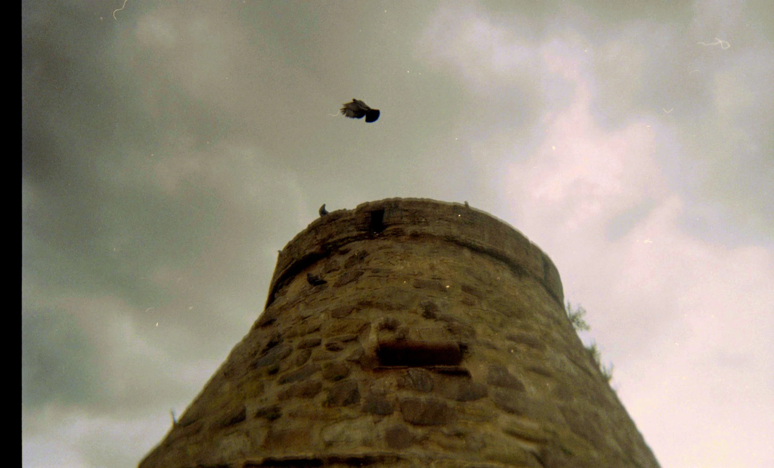 A low-angle view of a tall, rounded stone tower with a bird flying above it against a cloudy sky with faint lightning.