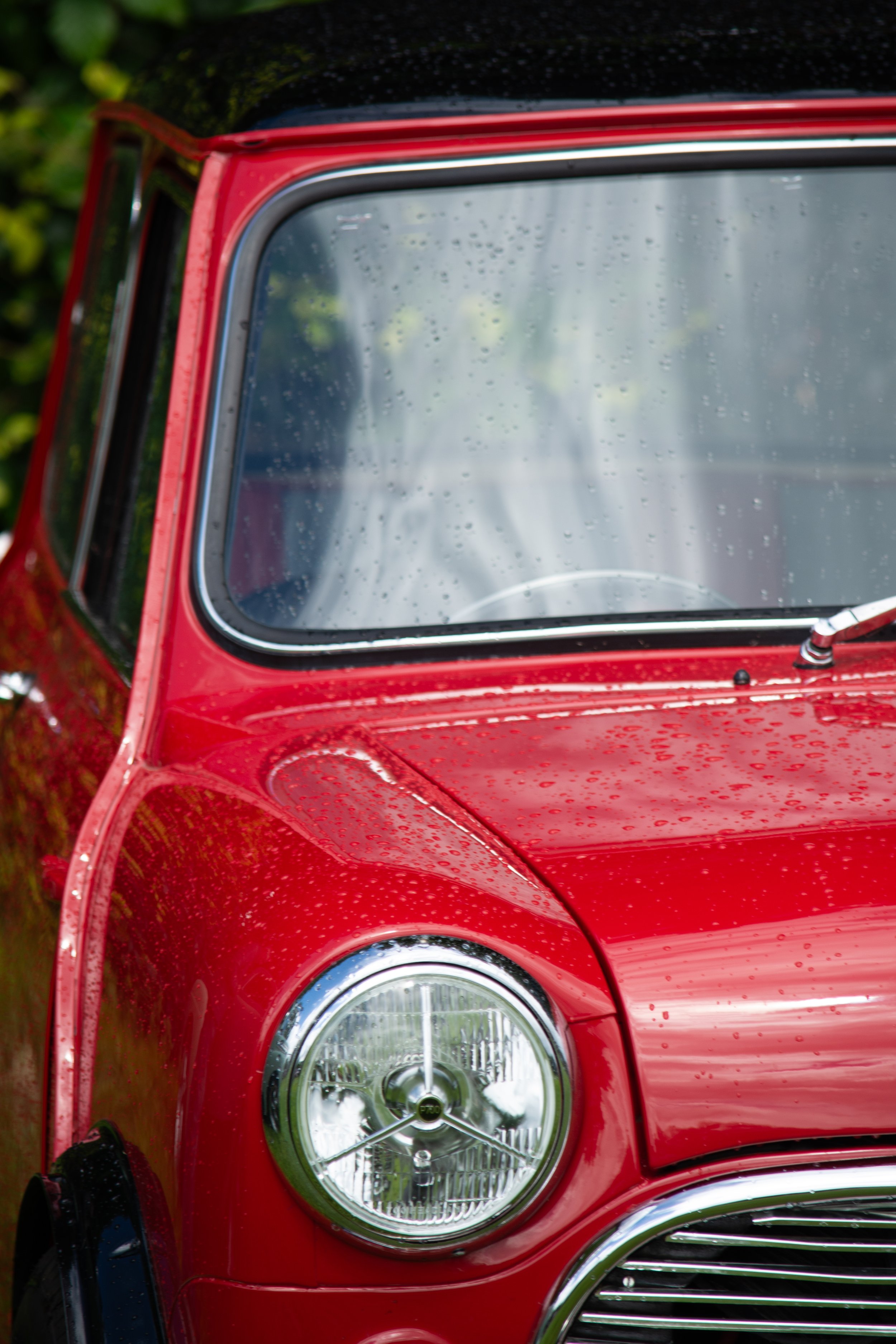 Close-up of the front part of a red classic car, showing the headlight, wheel, and windshield with rain droplets.