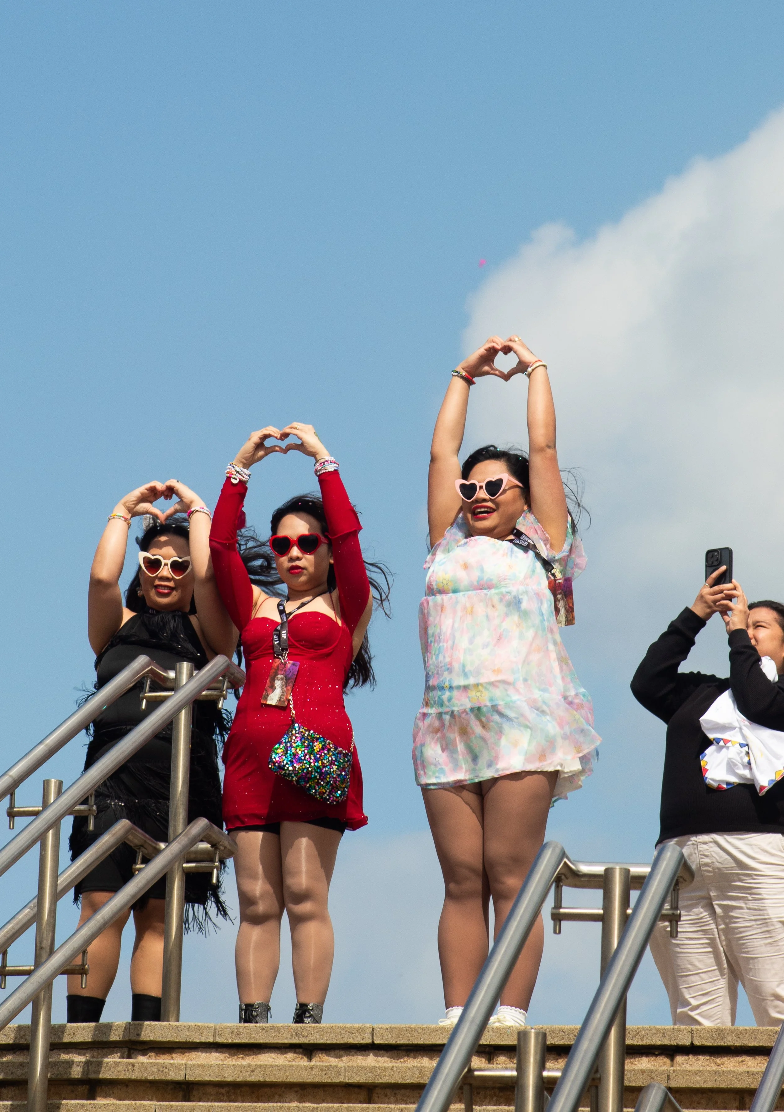 A group of young women standing on outdoor steps, making heart shapes with their hands, wearing colorful outfits and sunglasses, with a clear blue sky in the background.