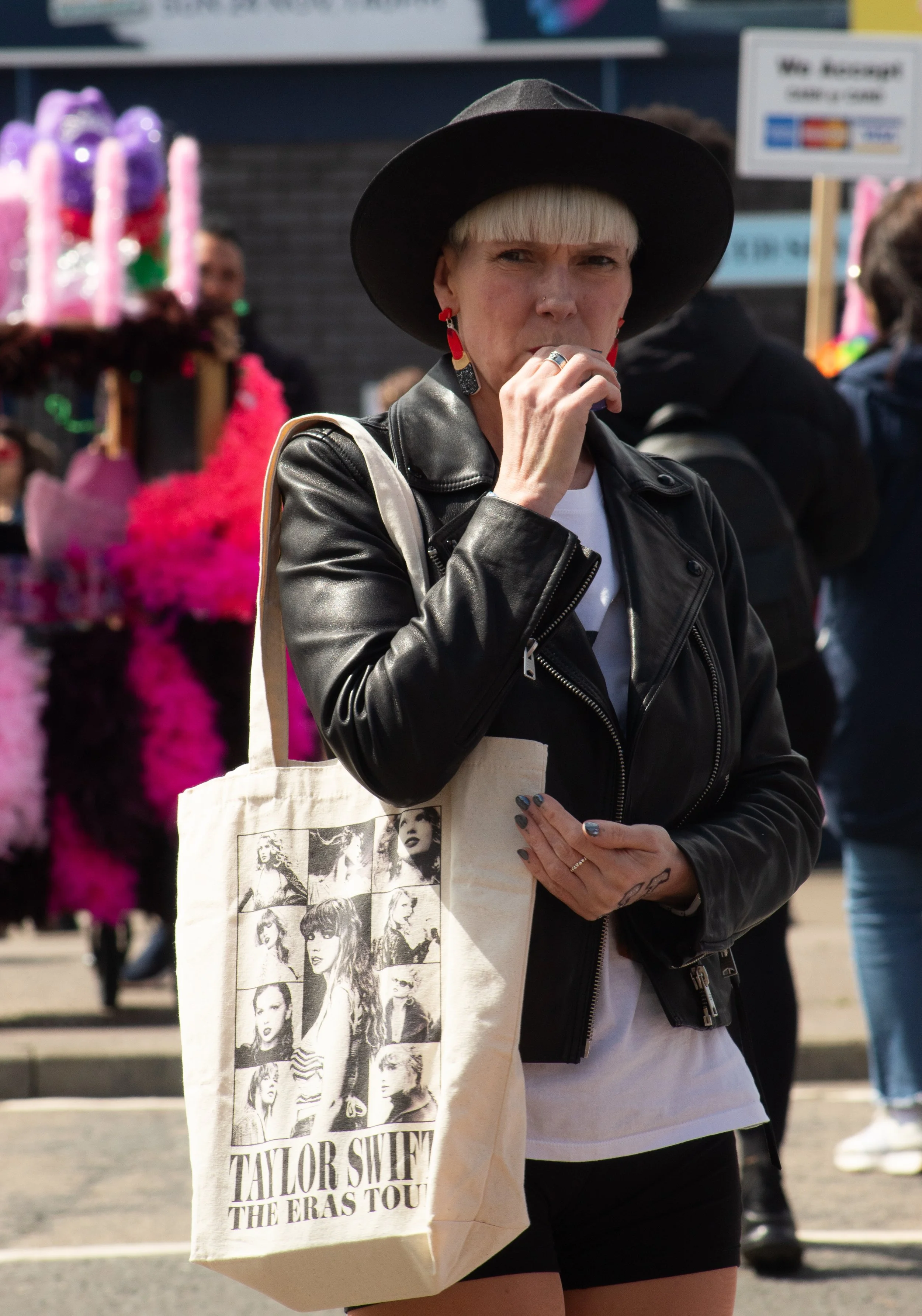 A woman wearing a black hat and leather jacket with a Taylor Swift tote bag at an outdoor event.