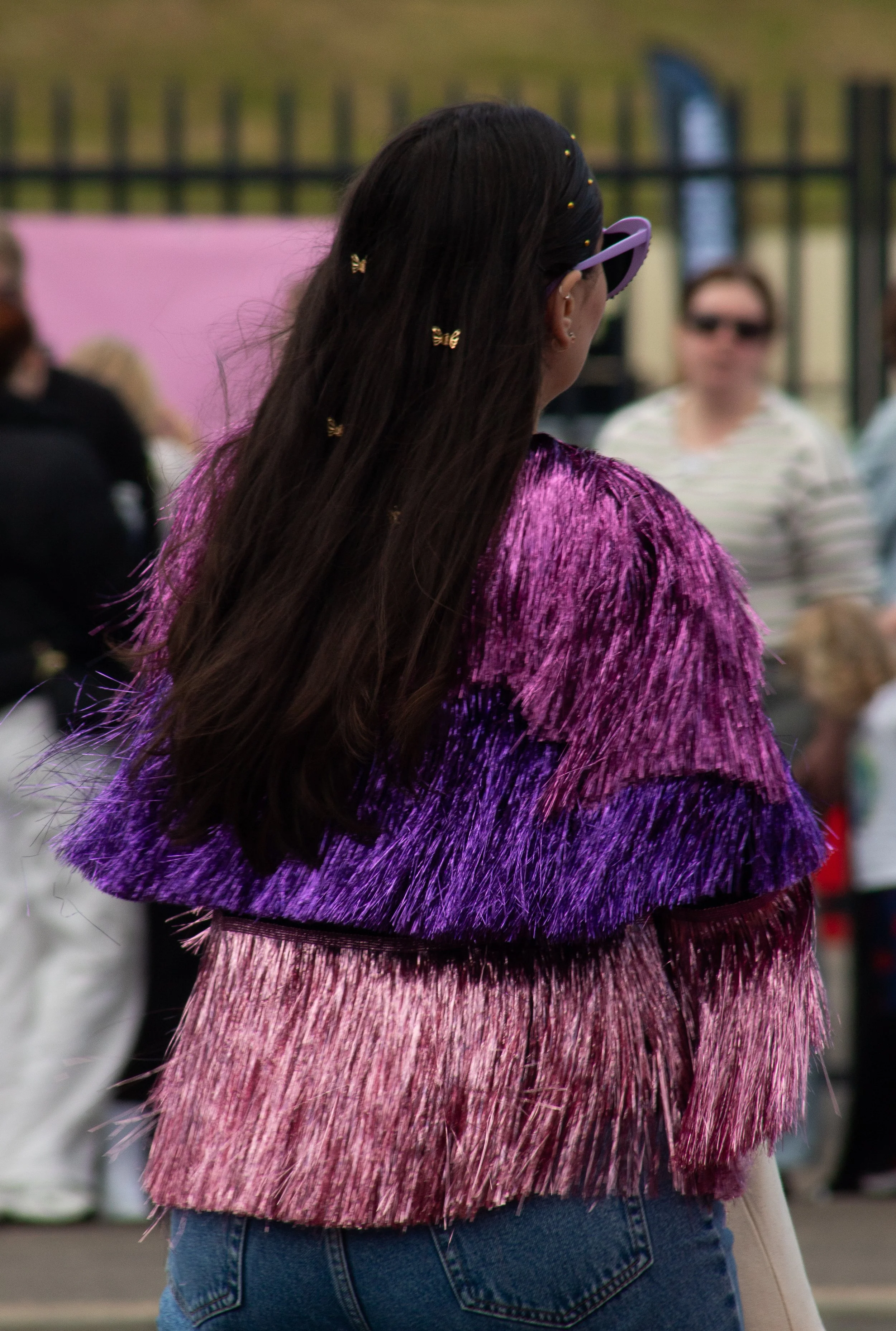 A woman with long dark hair adorned with small butterfly hair clips and butterfly earrings, wearing purple sunglasses and a vibrant, multi-colored feathered top in pink, purple, and red hues, standing outdoors with blurred people and a fence in the b