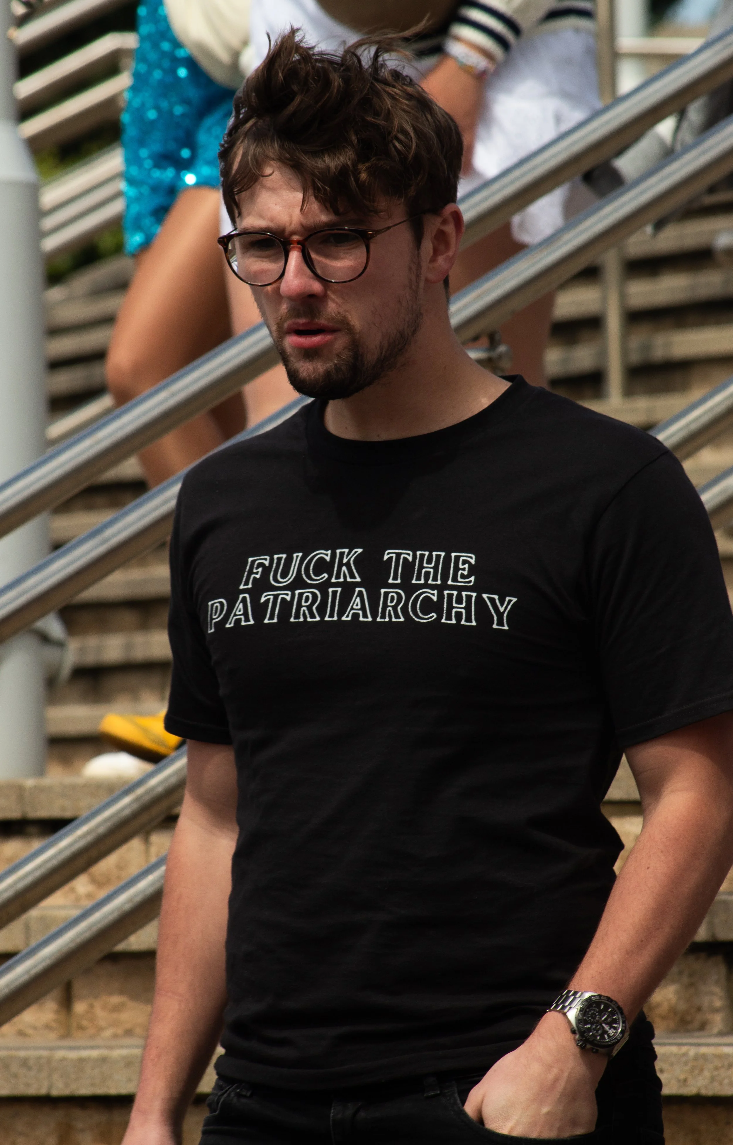 Young man wearing black T-shirt with a slogan, glasses, and a silver wristwatch, standing on outdoor stairs with metal railings, looking down with a serious expression.