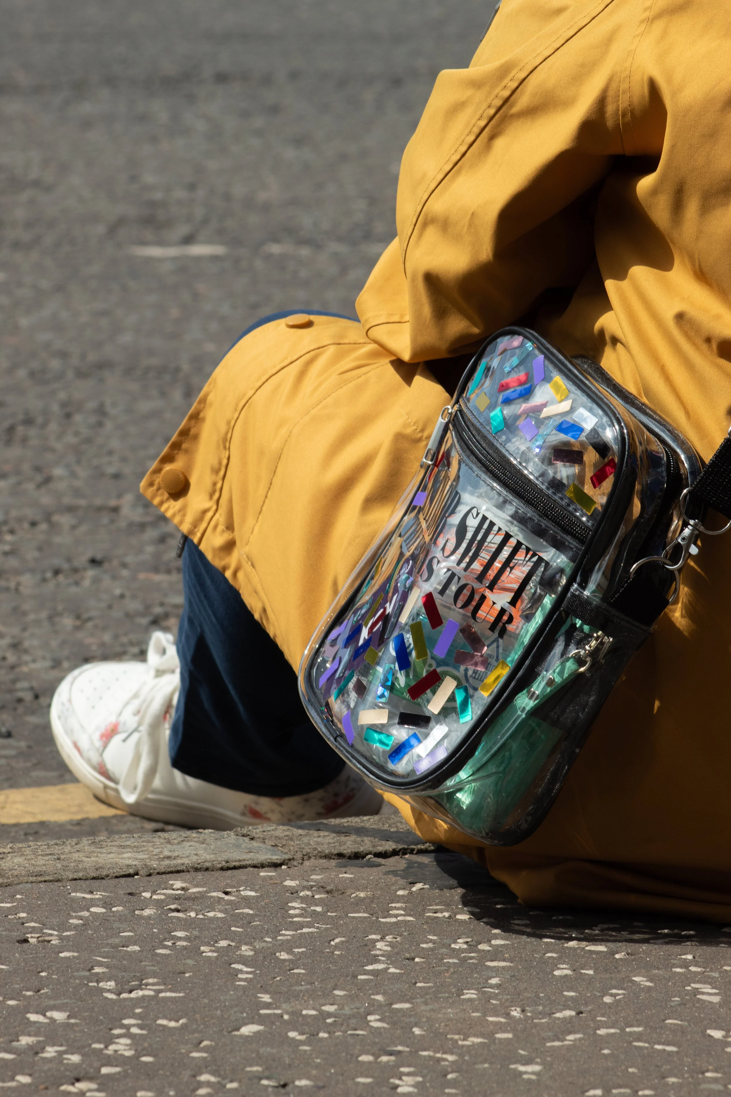 Person sitting on a curb wearing a mustard yellow jacket, dark pants, and white sneakers, holding a clear plastic purse filled with colorful confetti, with a gravel surface underneath.