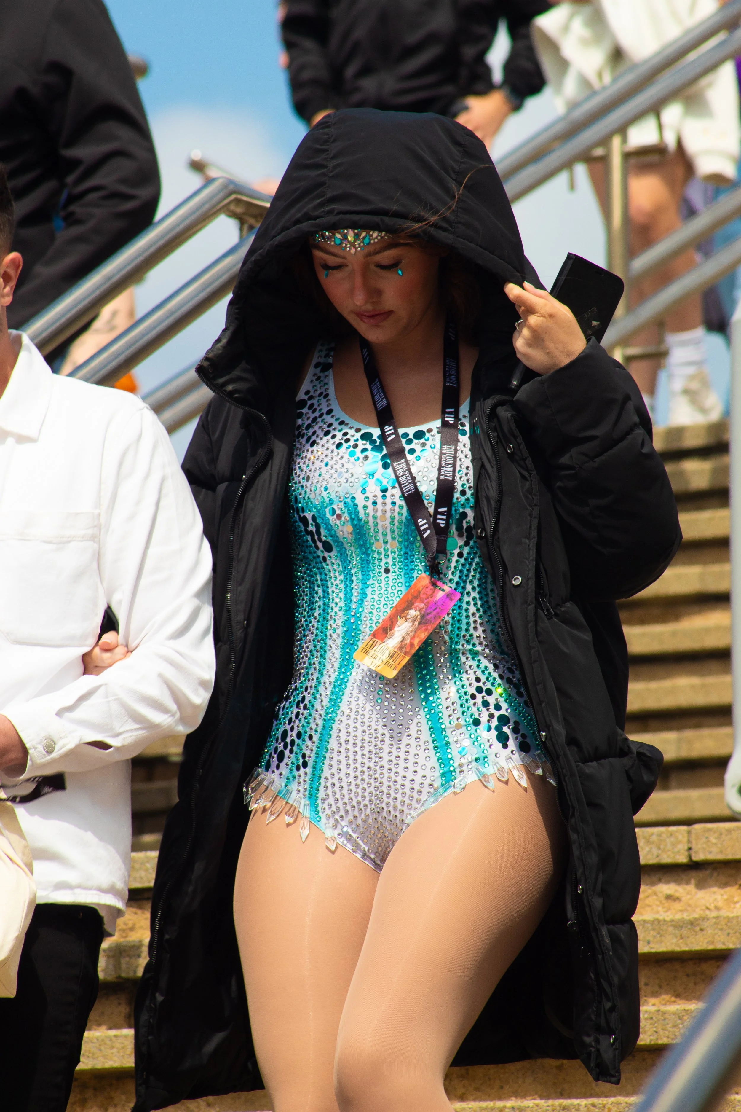 A woman wearing a sparkly, blue and silver dance costume and no pants, with a black jacket and a lanyard, walking down stairs outdoors.