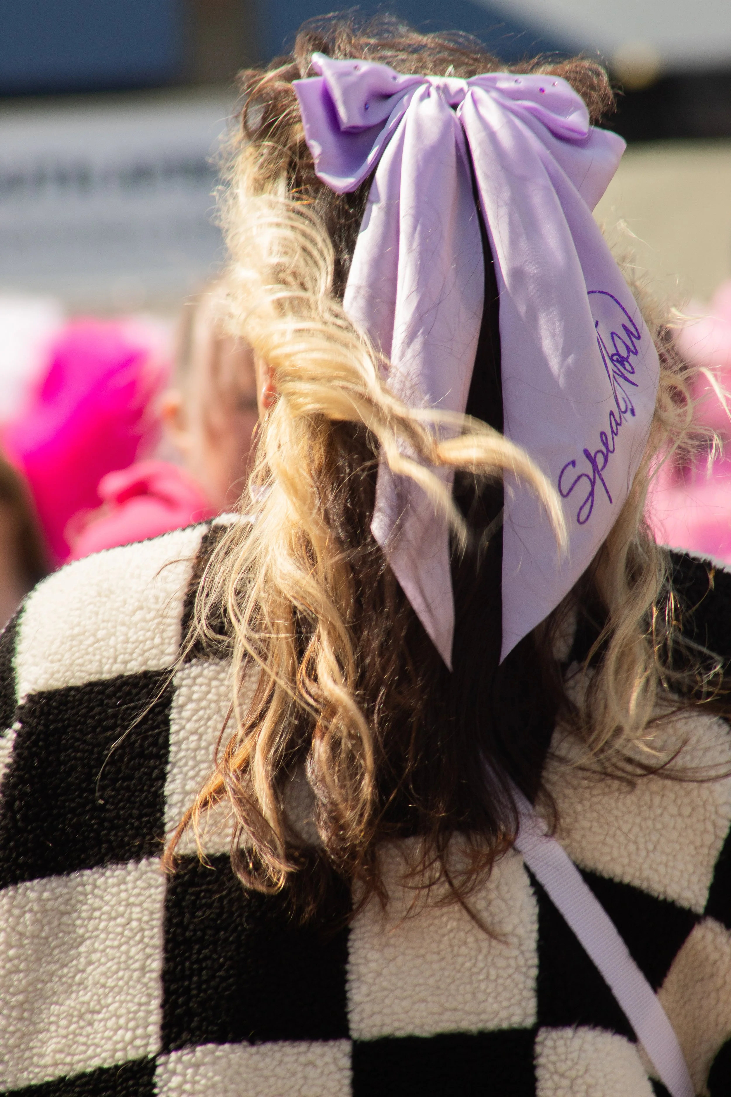 Close-up of a person wearing a large purple bow in their curly hair, with a light-colored, textured, striped sweater.