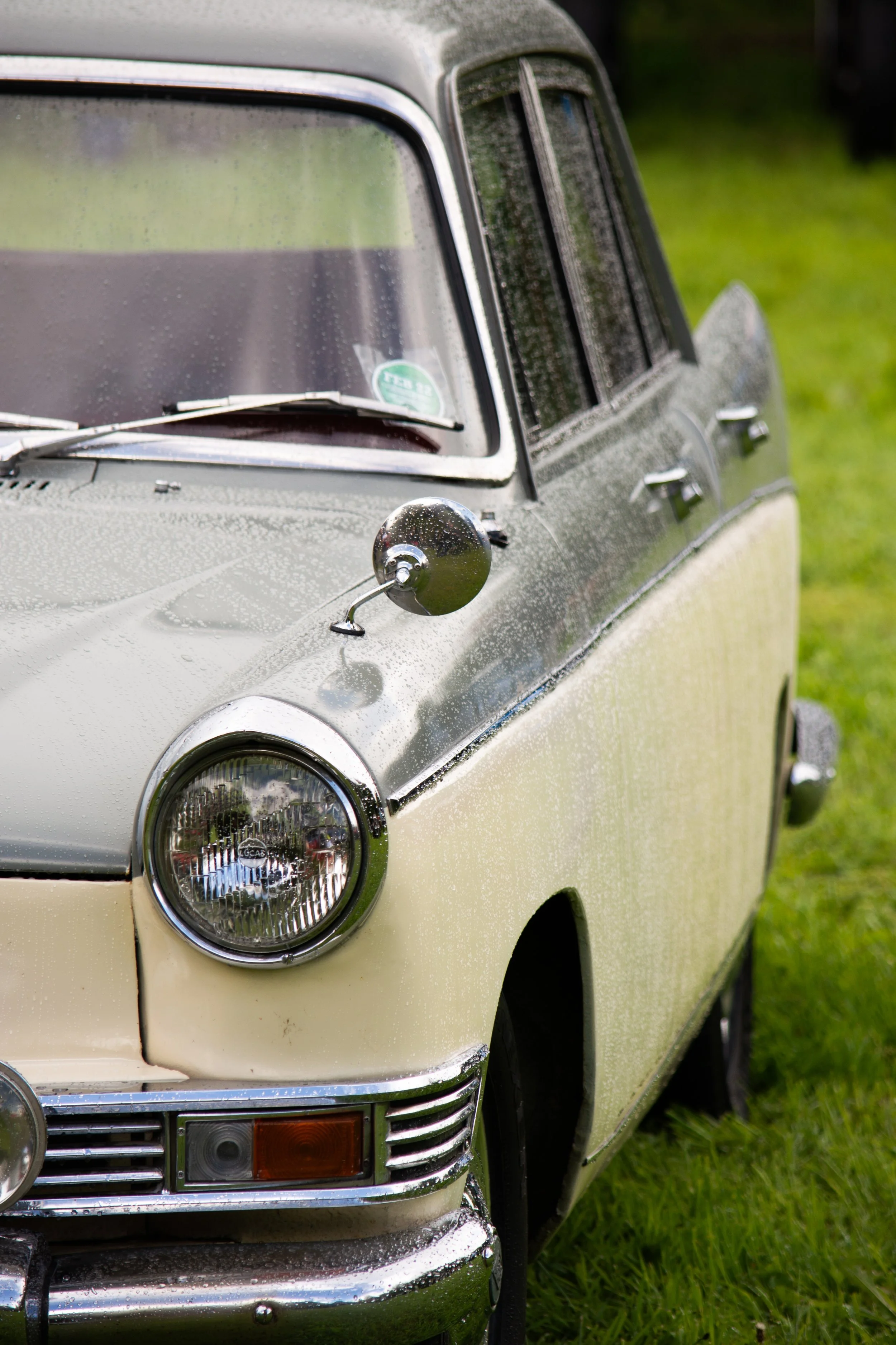 Close-up of the front portion of a vintage gray car with rain droplets on it, parked on grass.