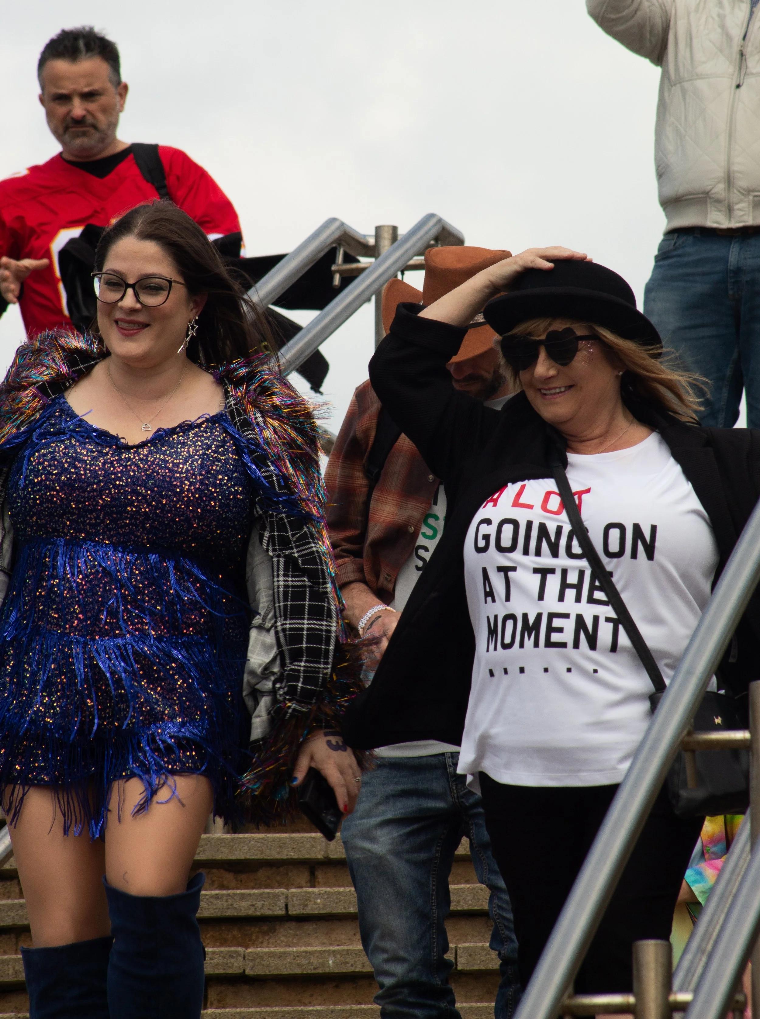 People standing and smiling on a set of outdoor steps, with some dressed casually and others in colorful outfits, including a woman in a sparkling dress and a woman with sunglasses and a hat.