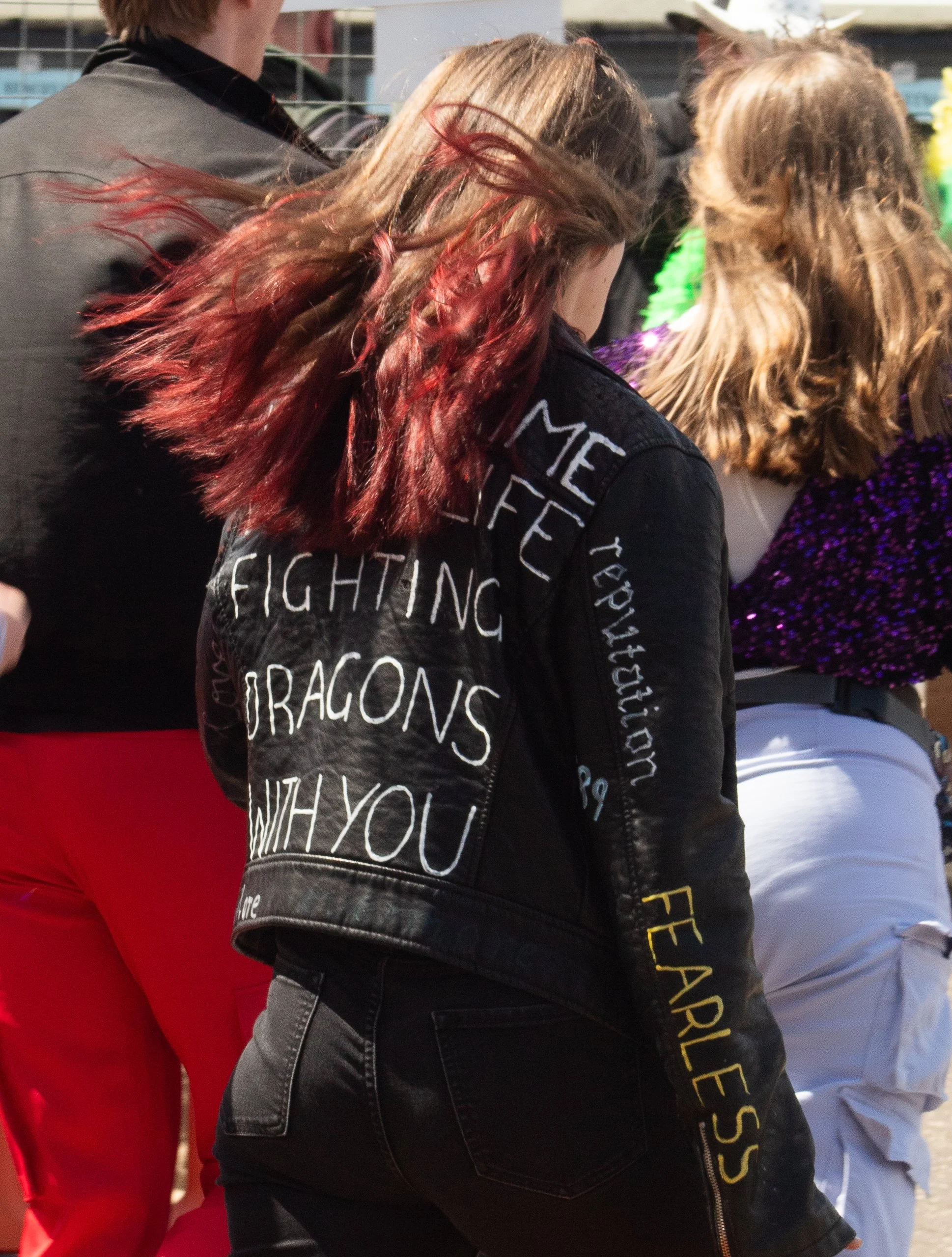 Close-up of a woman with red hair wearing a black jacket with text on the back, standing outdoors among other people, some in colorful clothing, at a public event.
