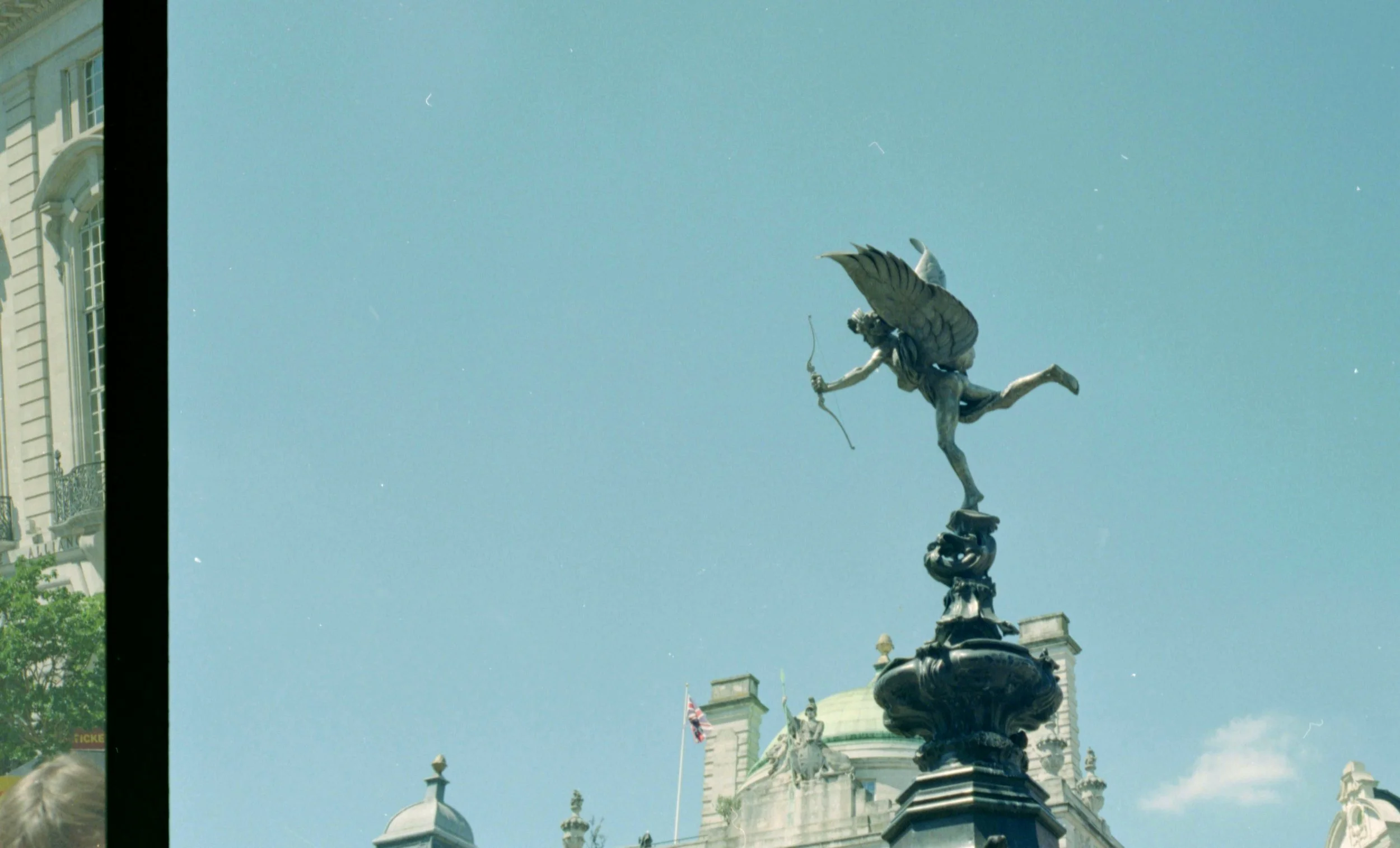 A statue of a figure with wings, holding a bow, atop a decorative pedestal, set against a blue sky and historic building with ornate architecture and flagpoles.