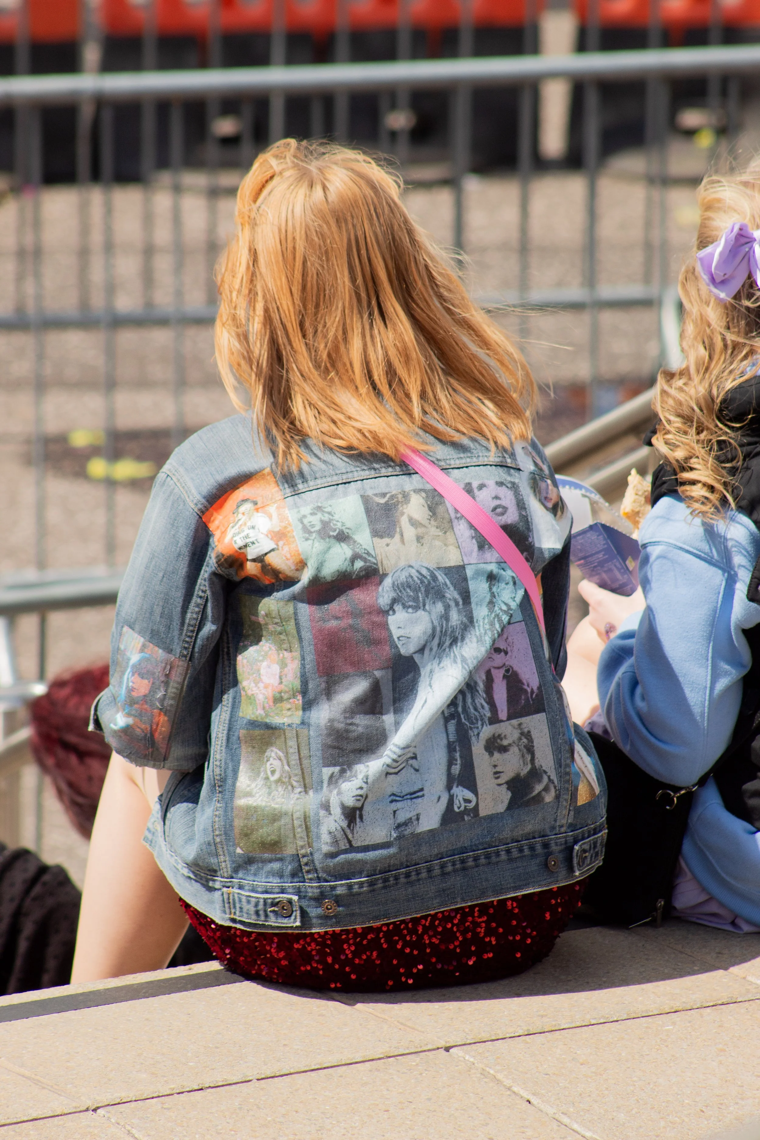 Back view of a girl with shoulder-length red hair wearing a denim jacket with pop art images on the back, sitting on a concrete ledge, with people and orange seats in the background.