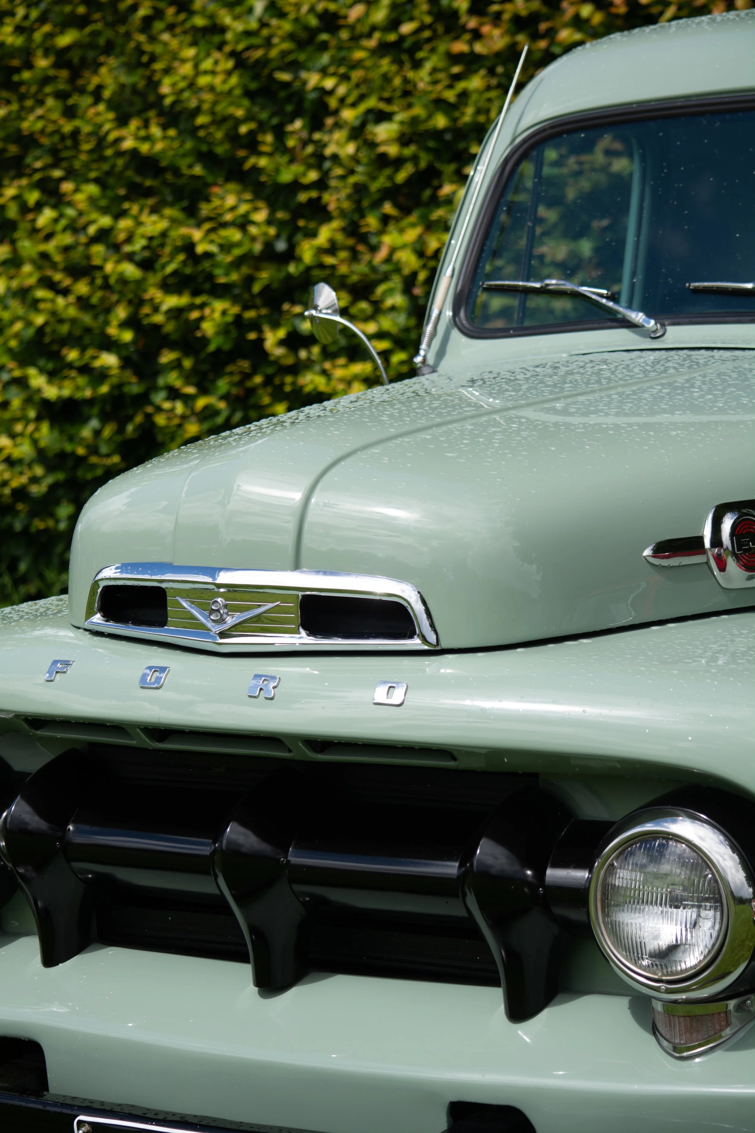 Close-up of the front of a vintage light green Ford car, showing the grille, headlights, and windshield with water droplets.
