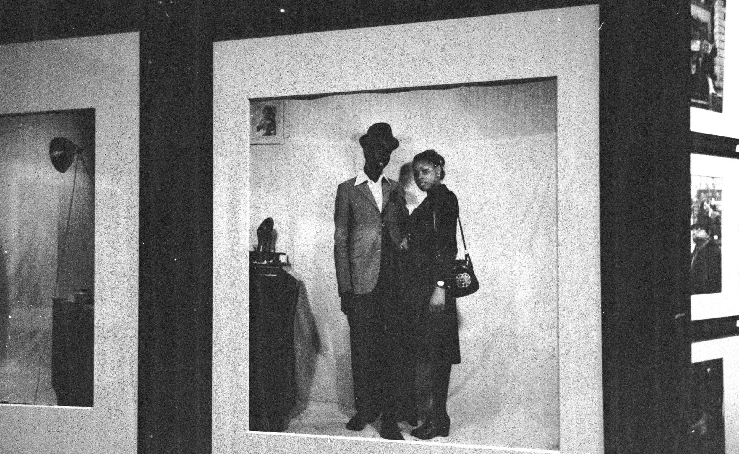 A black-and-white photograph of a young man and woman standing together inside a mirror exhibit. They are dressed stylishly, with the man in a suit and the woman in a coat or jacket. The mirror creates a reflection, making it appear as if they are standing in front of multiple lines or panels.