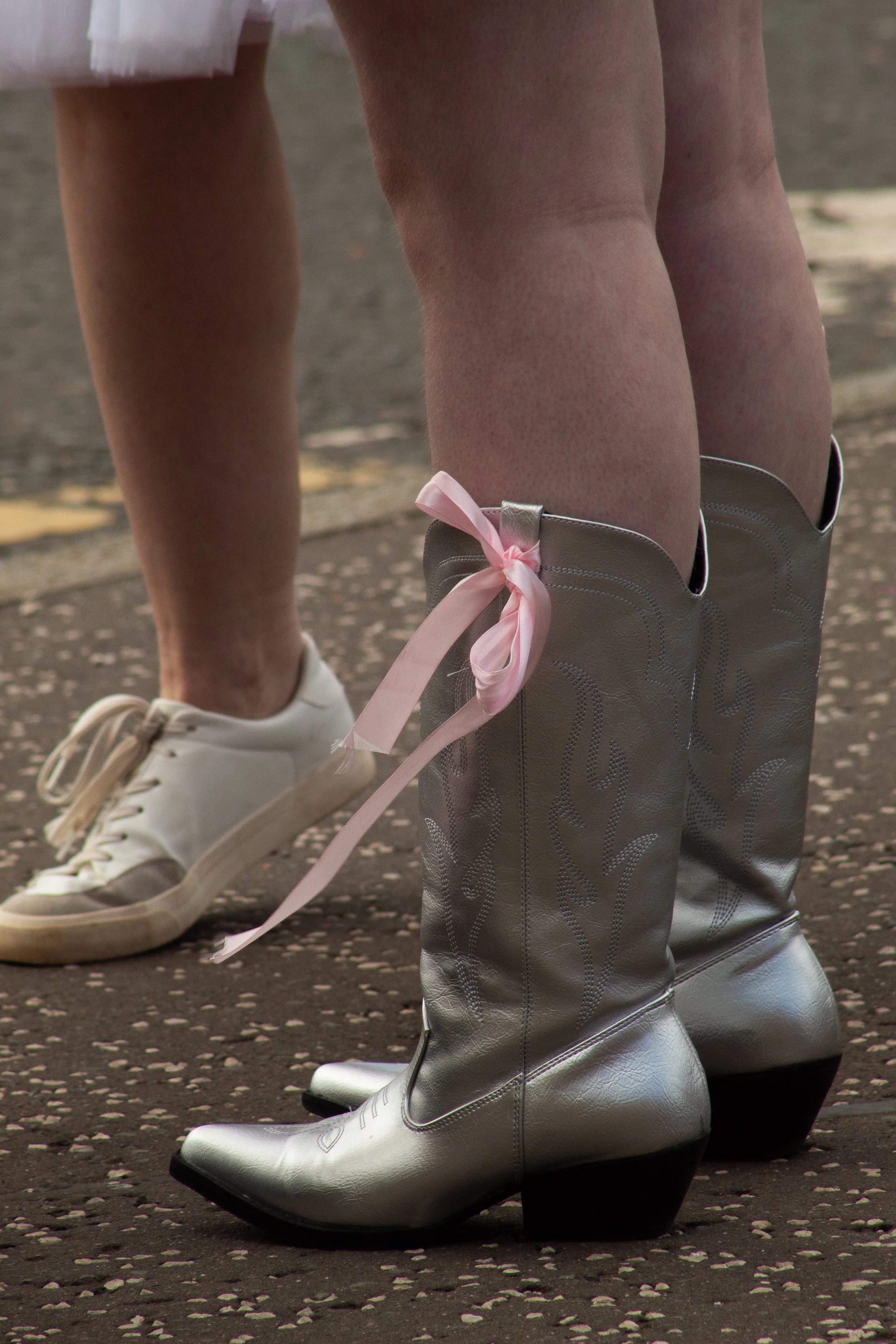 Close-up of someone wearing shiny silver ice skates with a pink ribbon tied to the boot, standing on an ice skating rink. Another person nearby is wearing white ice skates.