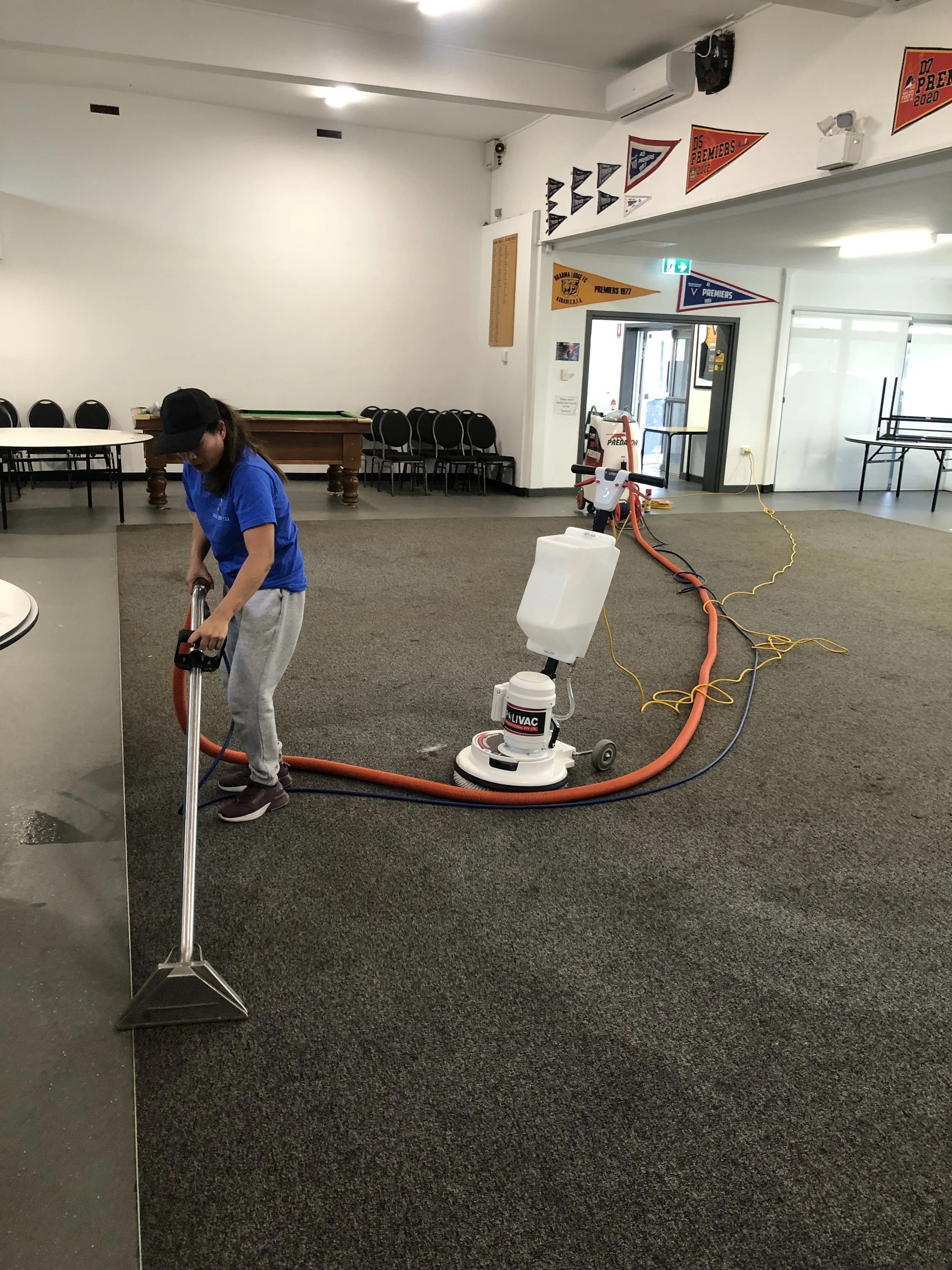 A person in a blue shirt and gray pants operating a carpet cleaning machine in an indoor room with some flags hanging on the wall.