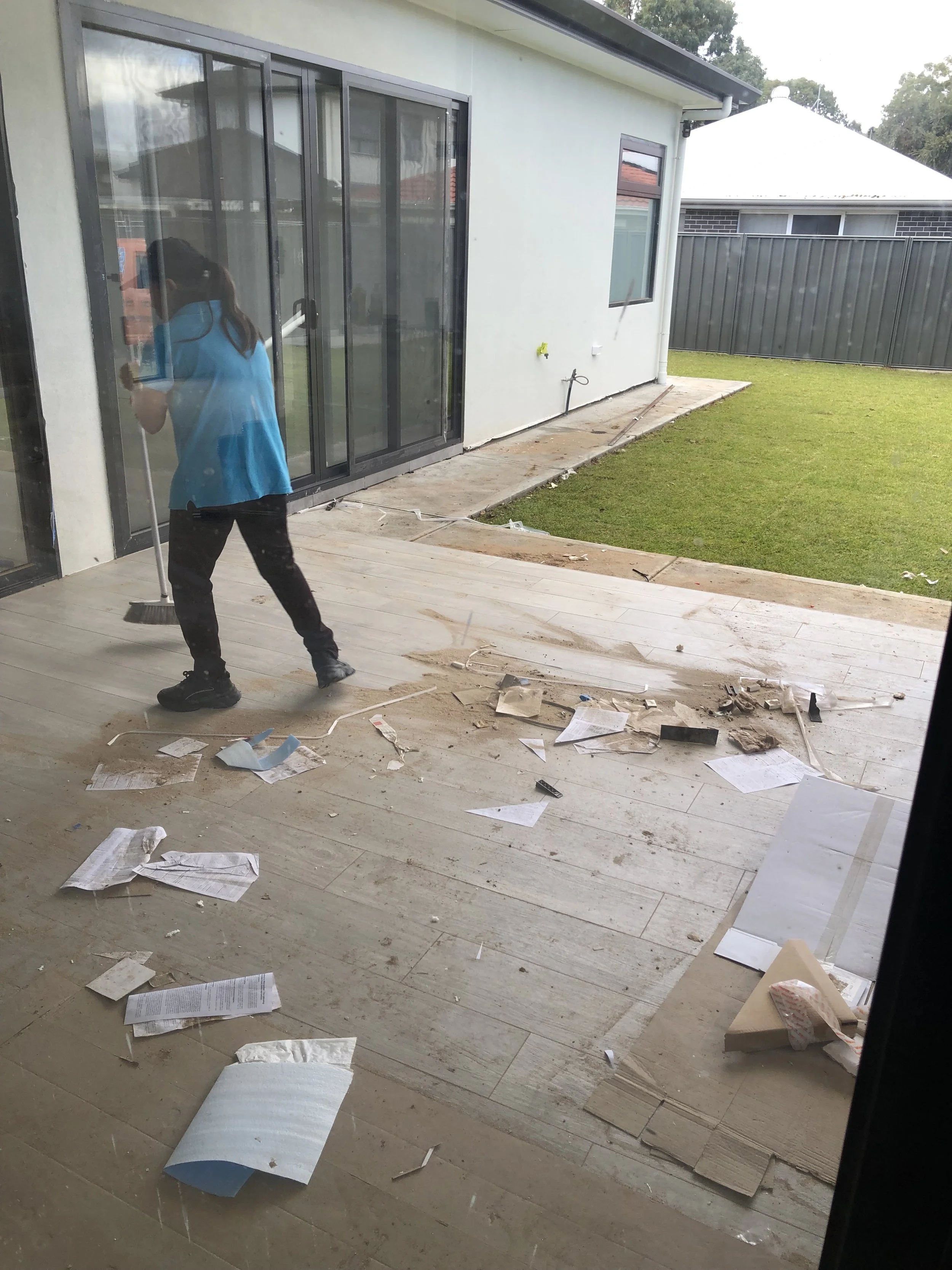 A person cleaning a glass door on a patio with scattered papers and debris on the floor, outside a house with a lawn and a neighboring house visible.