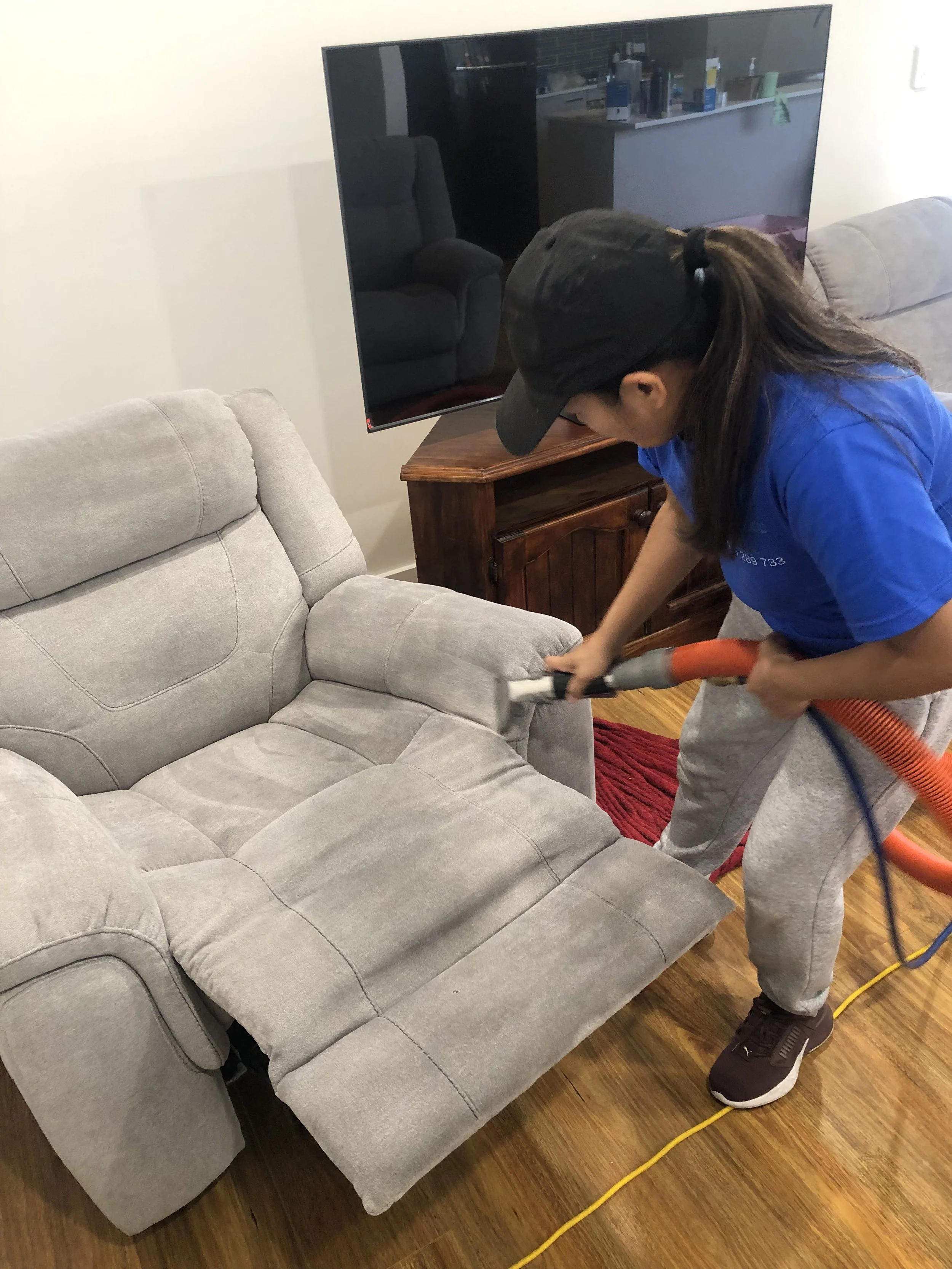 A person wearing a black cap and blue shirt vacuuming a gray upholstered sofa inside a living room.