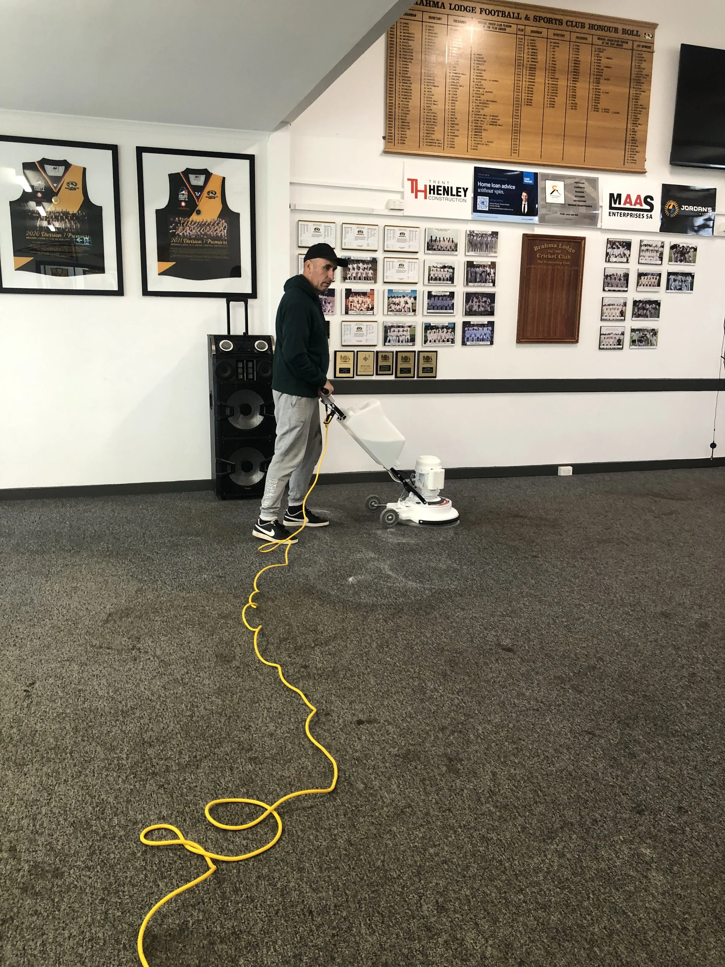 A man is cleaning a carpet with a floor cleaning machine in a room decorated with sports memorabilia and framed photographs.