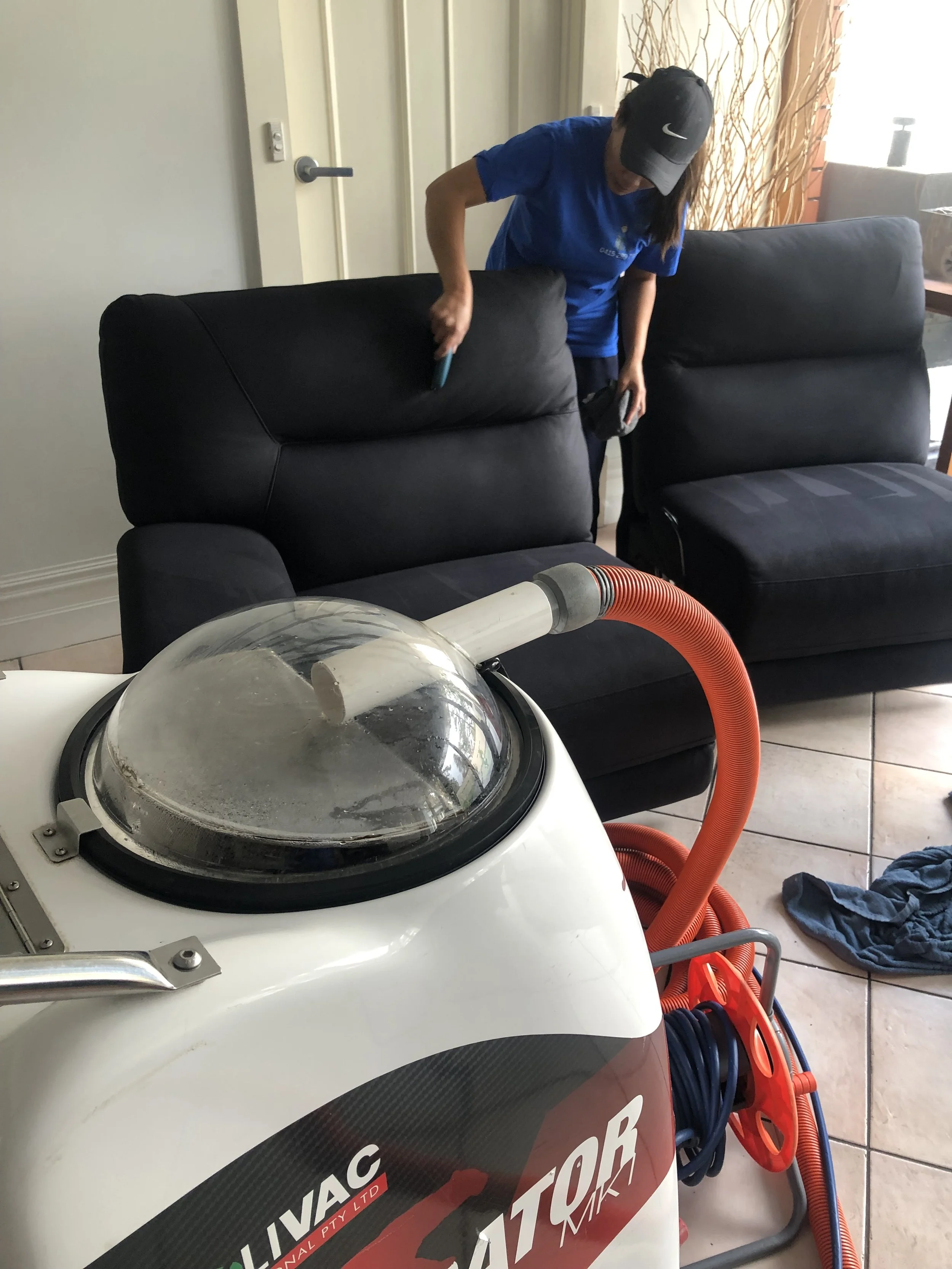 A woman dressed in blue cleaning a black sofa with a brush in a living room with tiled floor, with a carpet cleaning machine nearby.