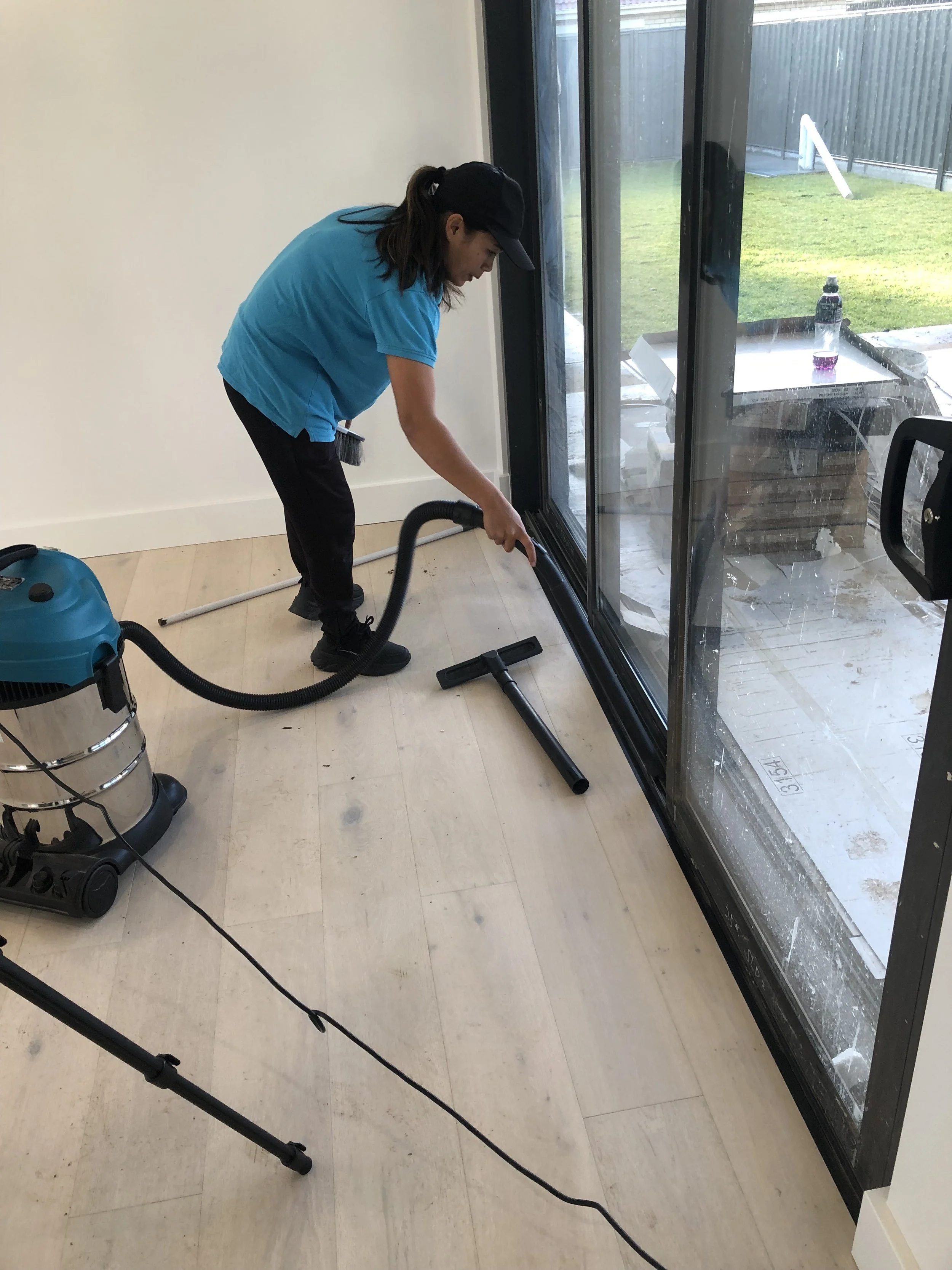 A woman vacuuming a light-colored wooden floor inside a room with large glass sliding doors leading to a patio and yard.