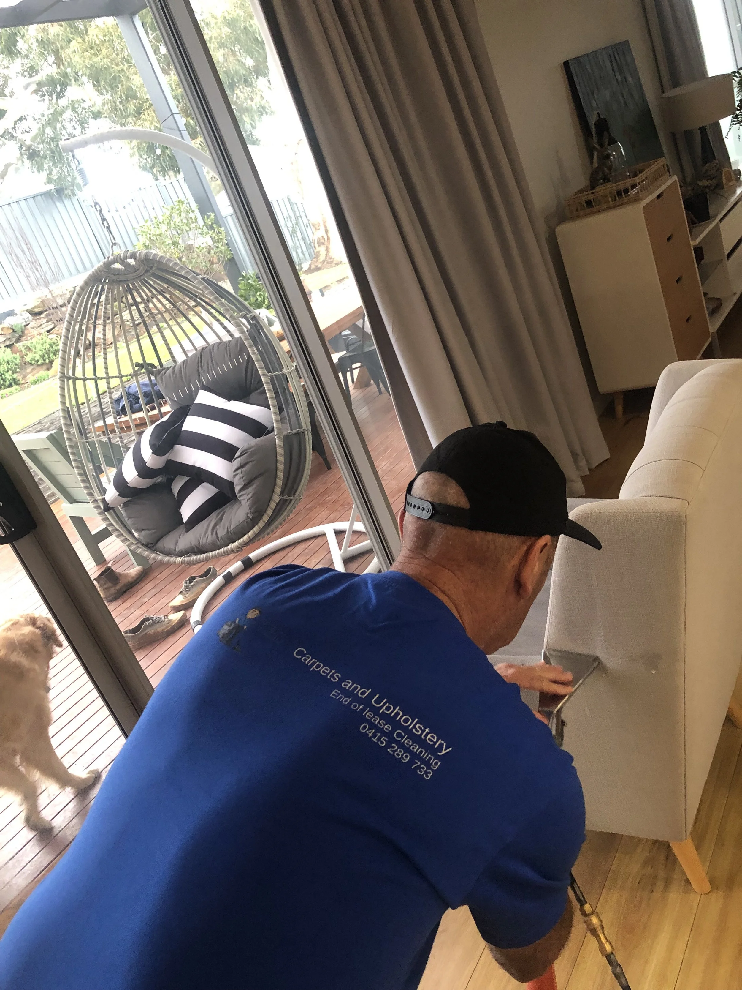Man cleaning or repairing the bottom part of a beige couch inside a living room, with sliding glass door leading to an outdoor patio with a hanging egg chair and a dog on the wooden floor.