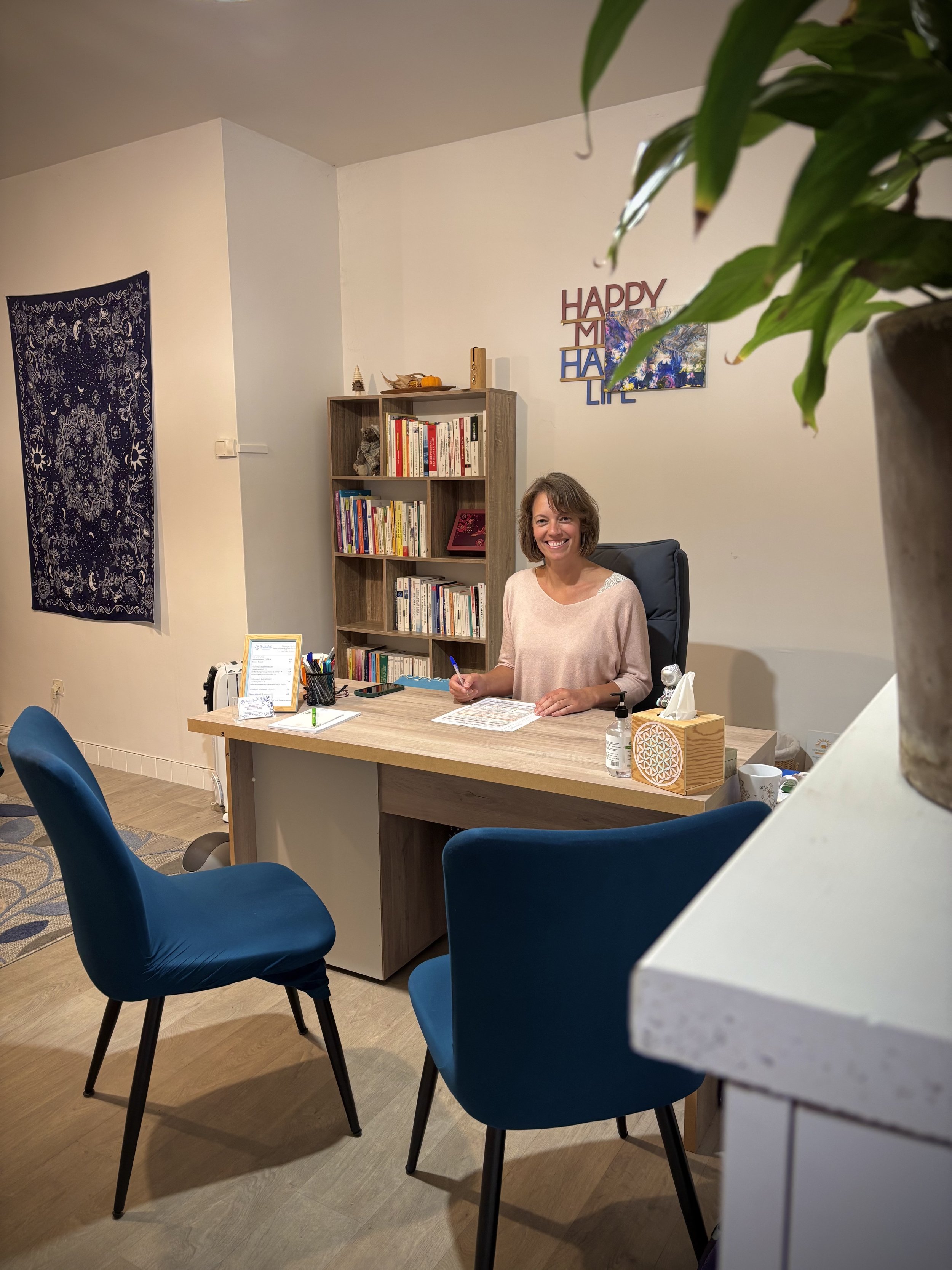 Une femme souriante - Charlotte Baele - assise derrière un bureau, dans un espace de bureau ou de consultation, avec une étagère de livres en arrière-plan et des décorations murales.