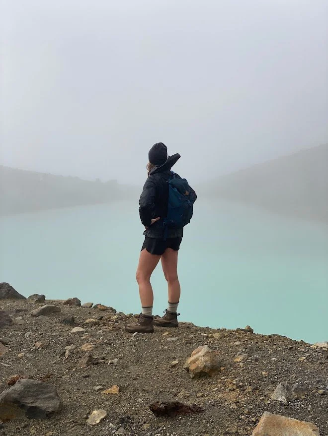 A hiker stands on rocky terrain near a body of water with a misty, foggy background, wearing a black jacket, shorts, hiking boots, and a blue backpack.