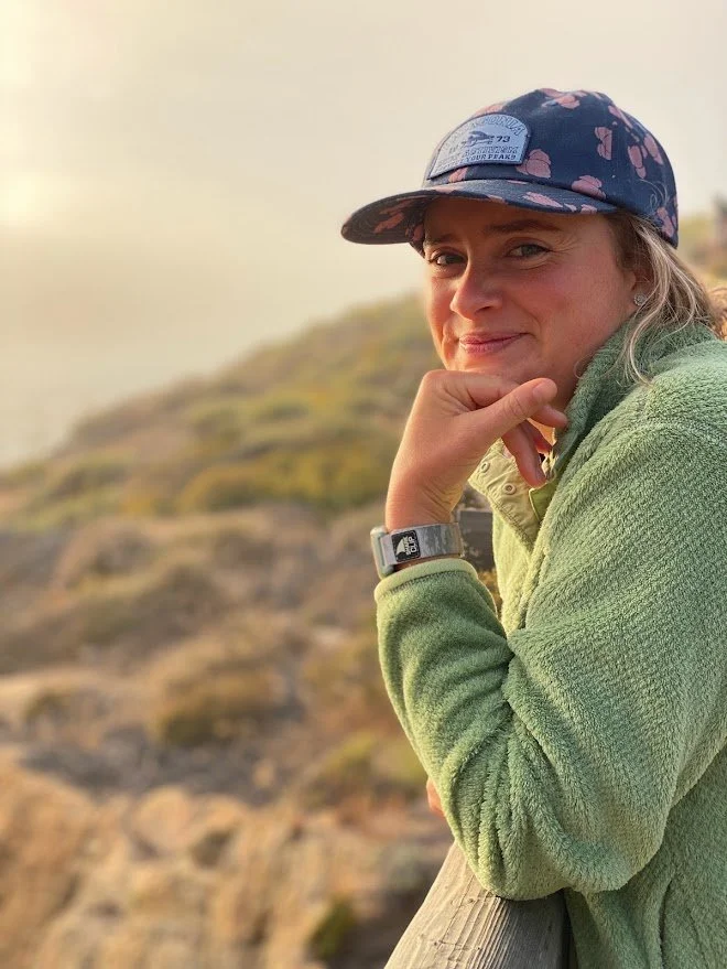 Person wearing a cap and green fleece sweater standing outdoors, smiling with a scenic blurred background.
