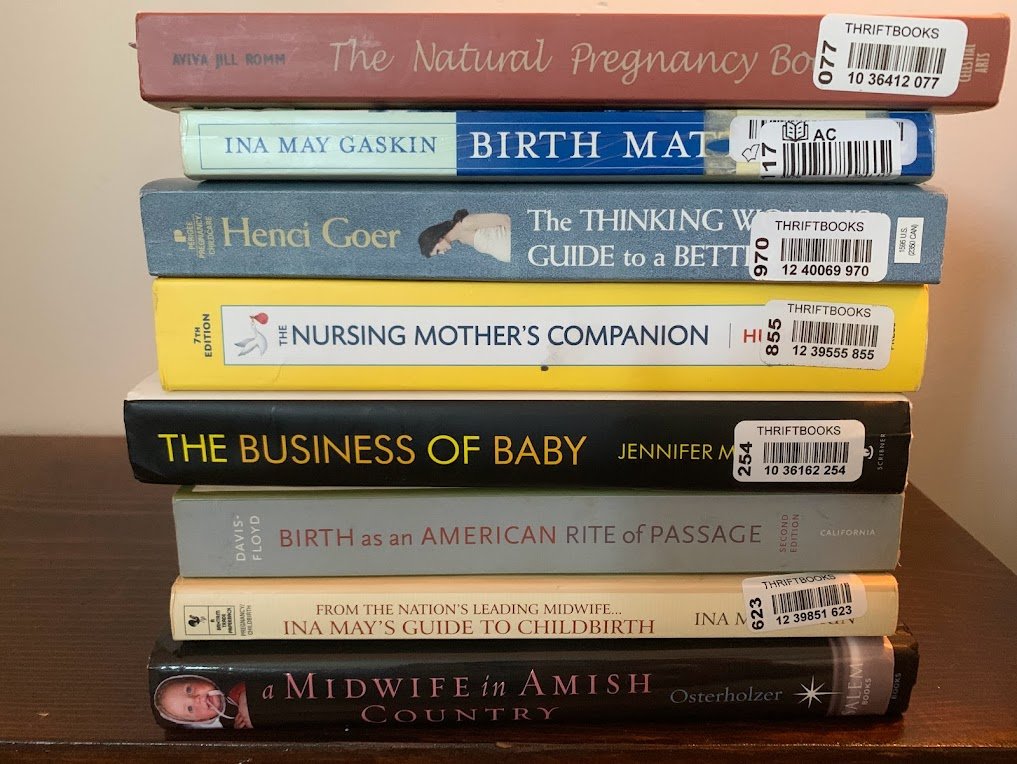 Stack of nine books on a wooden surface. Titles include 'The Natural Pregnancy Book,' 'Birth Mat,' 'The Thinking Woman's Guide to a Better Birth,' 'The Nursing Mother's Companion,' 'The Business of Baby,' 'Birth as an American Rite of Passage,' 'From the Nation's Leading Midwife...Ina May's Guide to Childbirth,' and 'A Midwife in Amish Country.'